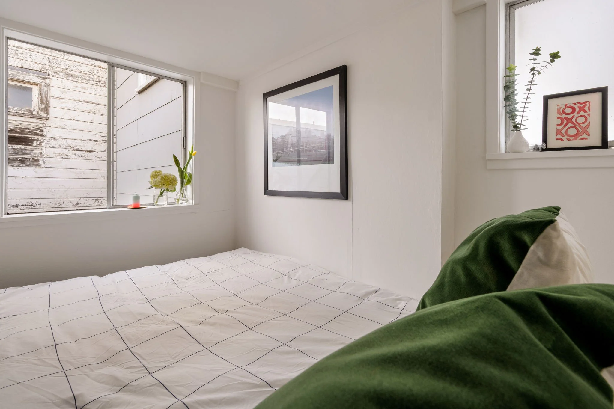 A cozy white bedroom with a window, decorated with framed artwork and potted plants, with a bed covered in a white grid-patterned duvet and green pillows.