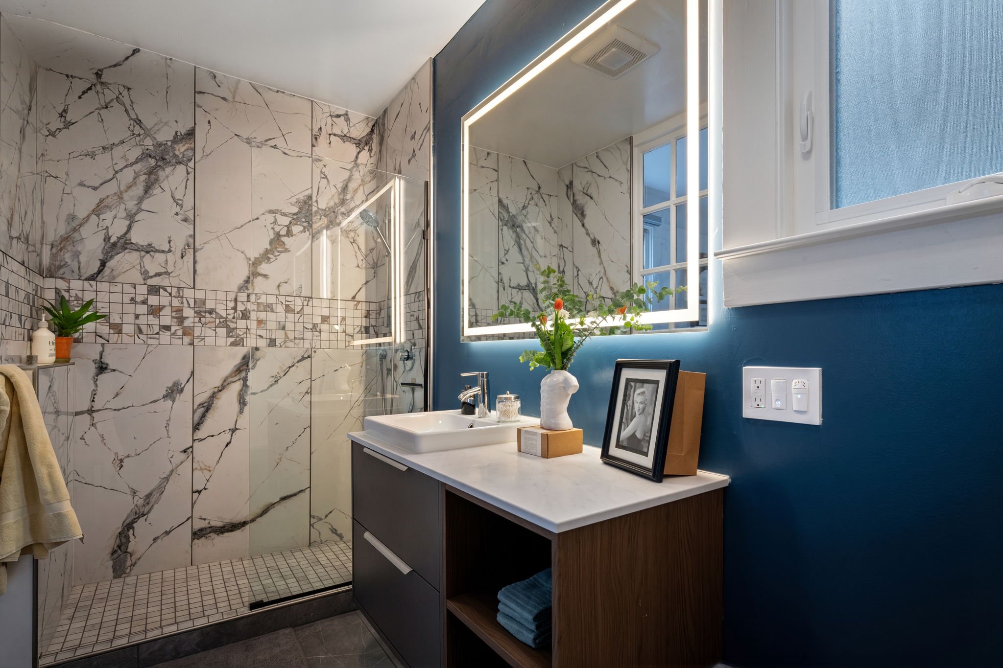 Modern bathroom with marble tiles, a small shower area with a glass door, a sink vanity with framed photos and decor, a window with a blue wall, and decorative plants.
