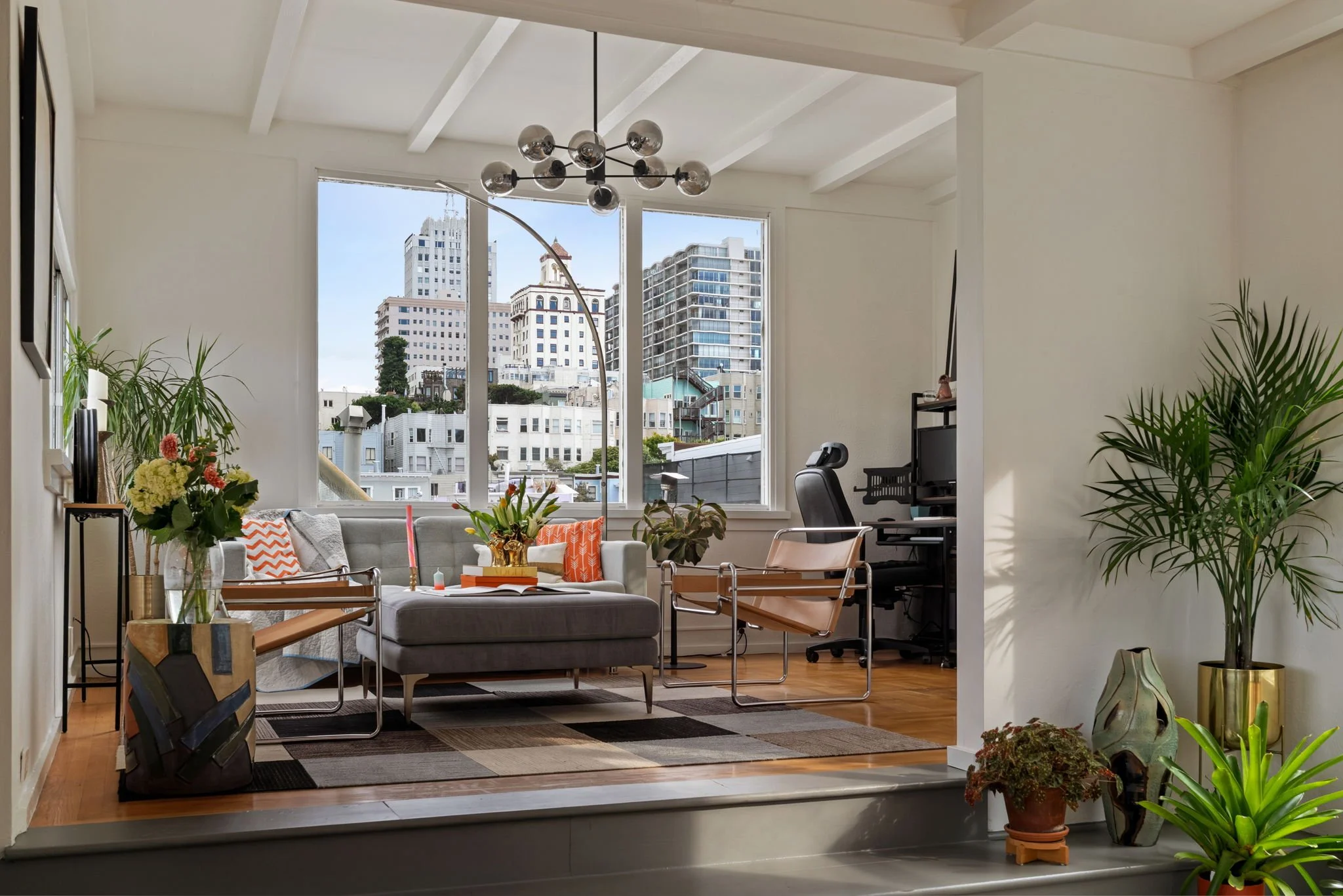 Interior of a modern apartment living room with large windows showing city buildings, a gray sofa with orange and white pillows, various plants, a black desk with a computer, and a contemporary chandelier overhead.