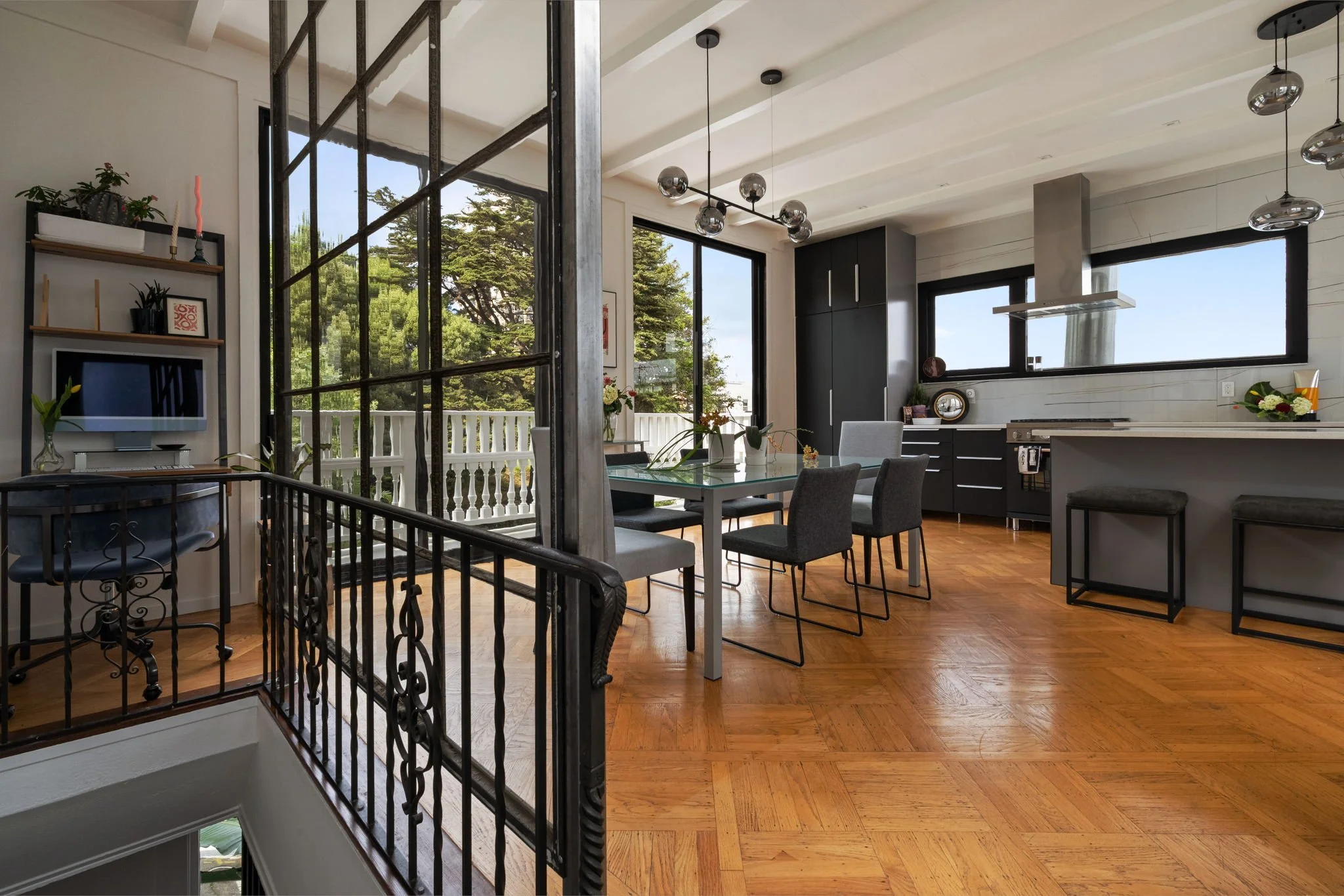 Modern kitchen and dining area with large windows showing trees outside. Features black cabinets, a white table with gray chairs, and hanging spherical pendant lights.