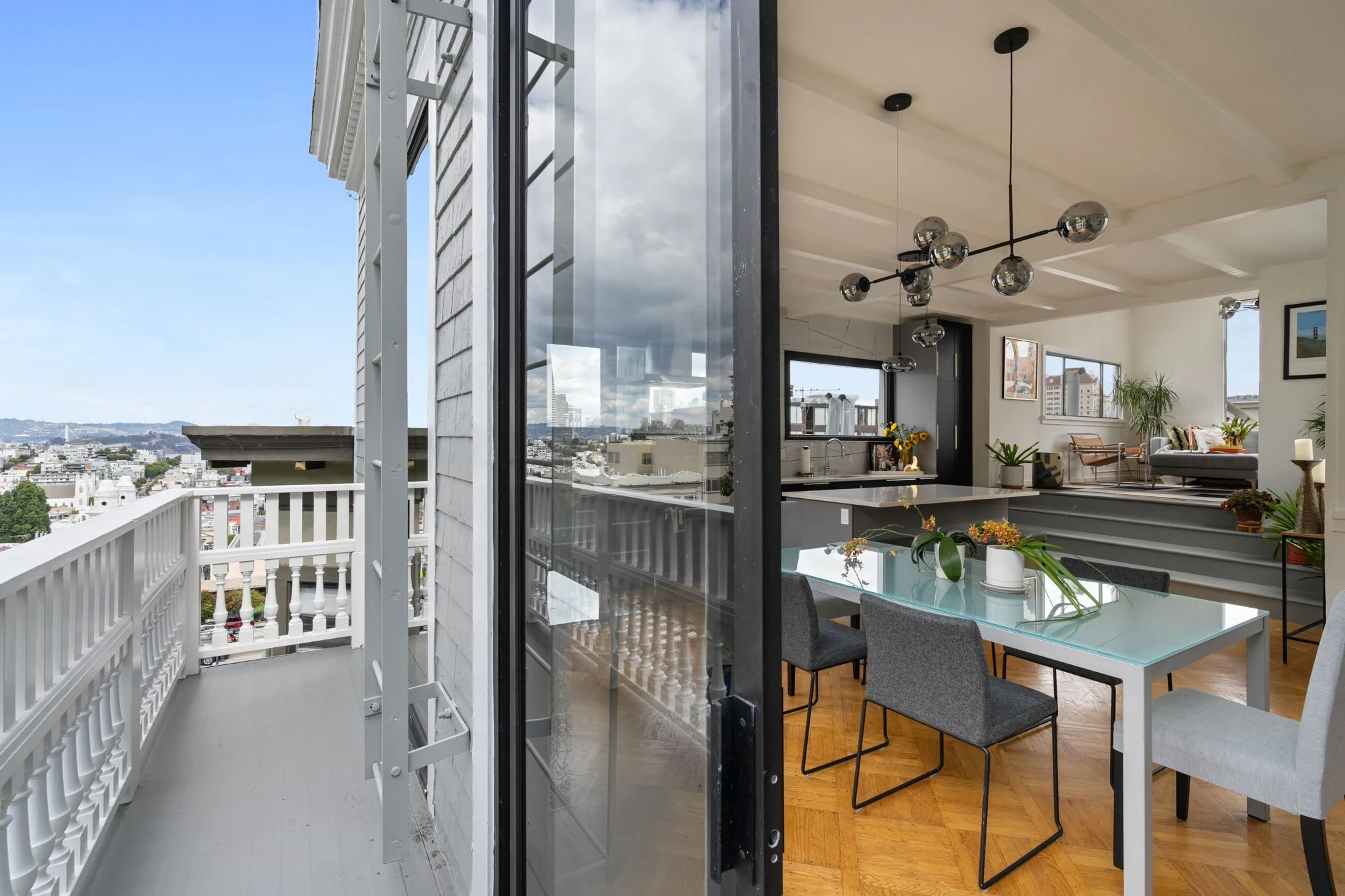 A modern apartment interior with a view of the city from the balcony. The living area has a dining table with four chairs, plants, wall art, and a cozy seating area with a sofa and chair. Ceiling lights hang from the ceiling, and large windows and sl