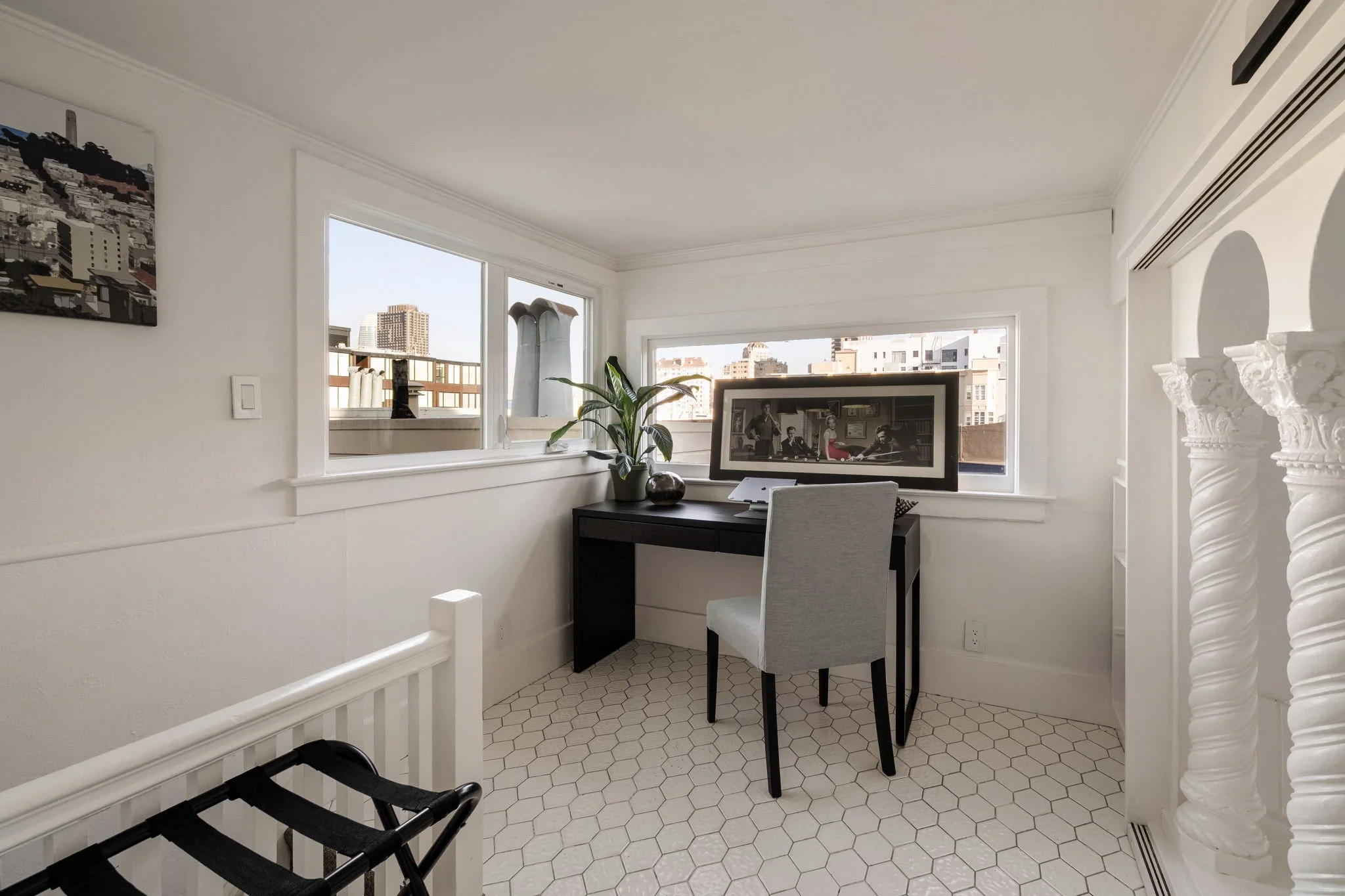 A bright corner of a room with two large windows showing city buildings outside. Inside, there is a white desk with a framed photo, a potted plant, and a computer monitor. A grey upholstered chair is in front of the desk. On the left, part of a black