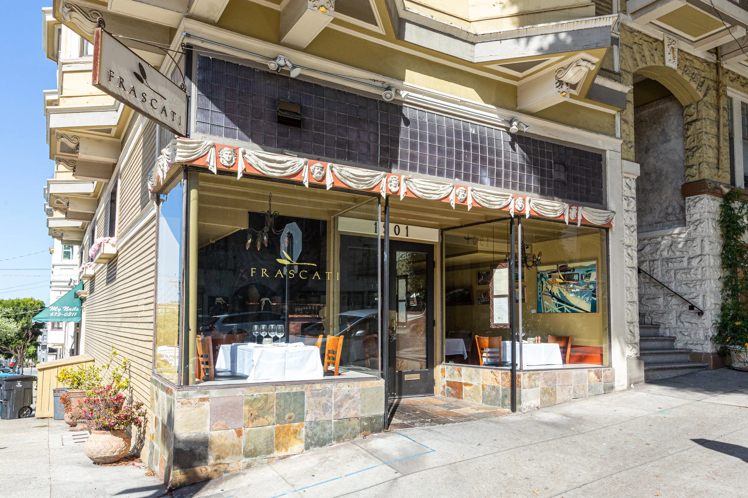 The image shows a corner of a restaurant with large glass windows and outdoor seating. The restaurant name 'FRASCATI' is visible on the window. The building has decorative trim and a sign that reads 'FRASCATI' hanging above. There are some chairs and