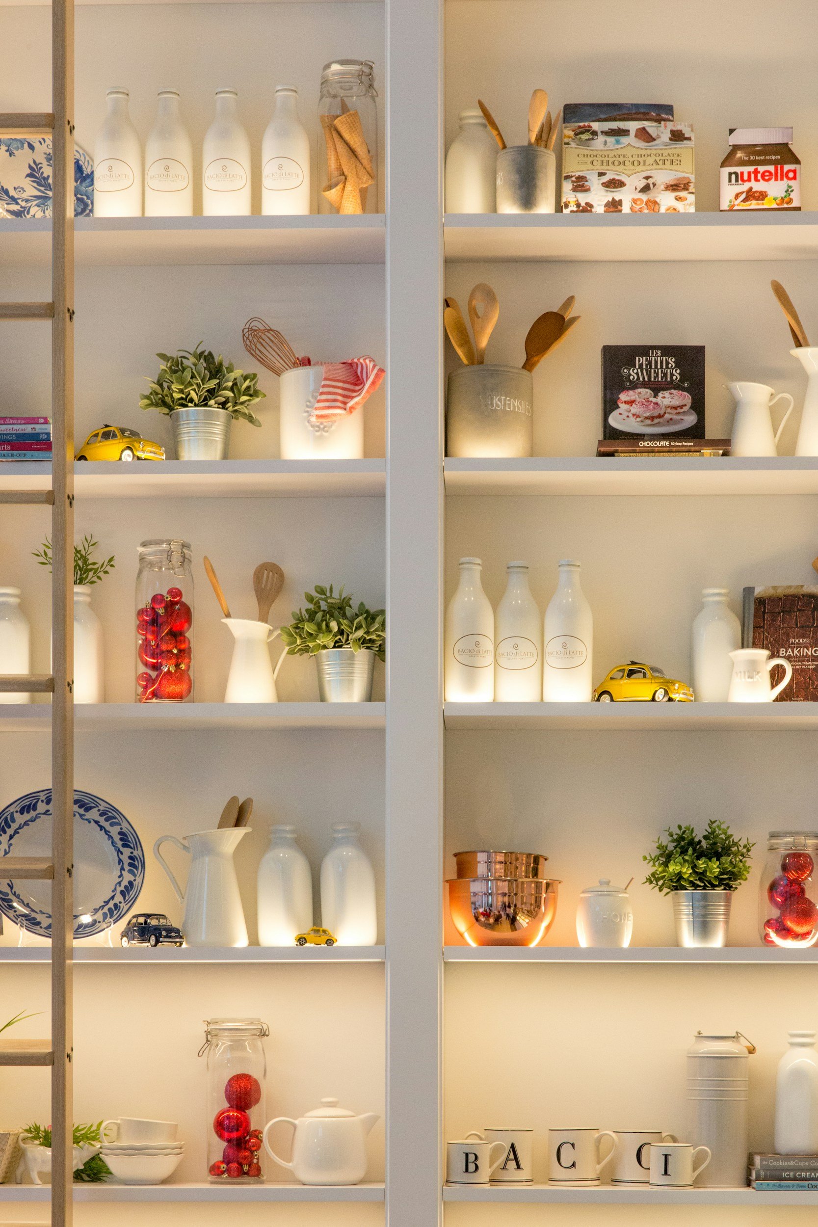 White kitchen shelves filled with decorative items including white bottles, small toy cars, potted plants, glass jars with red ornaments, cups labeled with letters, and cookbooks.