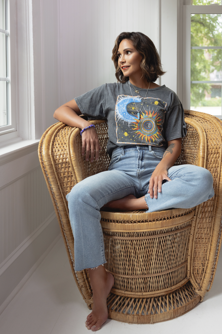 A woman with short dark hair and light skin relaxing in a wicker chair near a window with greenery outside, wearing a graphic T-shirt and jeans.