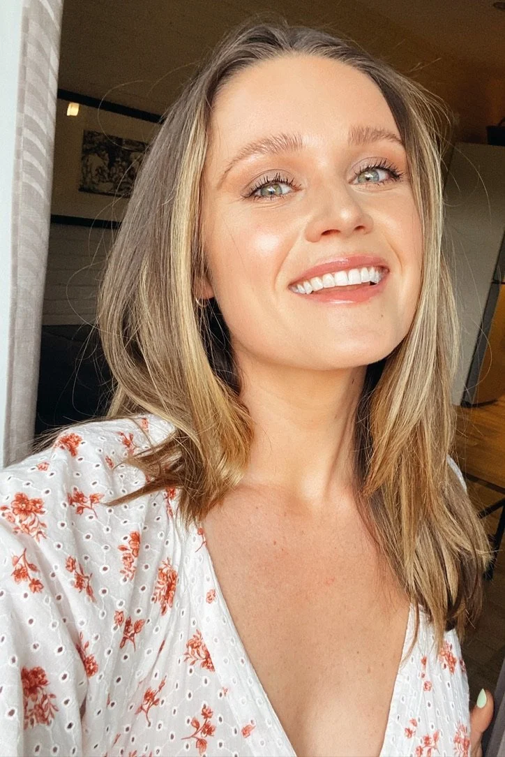 A smiling young woman with shoulder-length blonde hair and blue eyes, wearing a white blouse with red floral patterns, standing indoors.