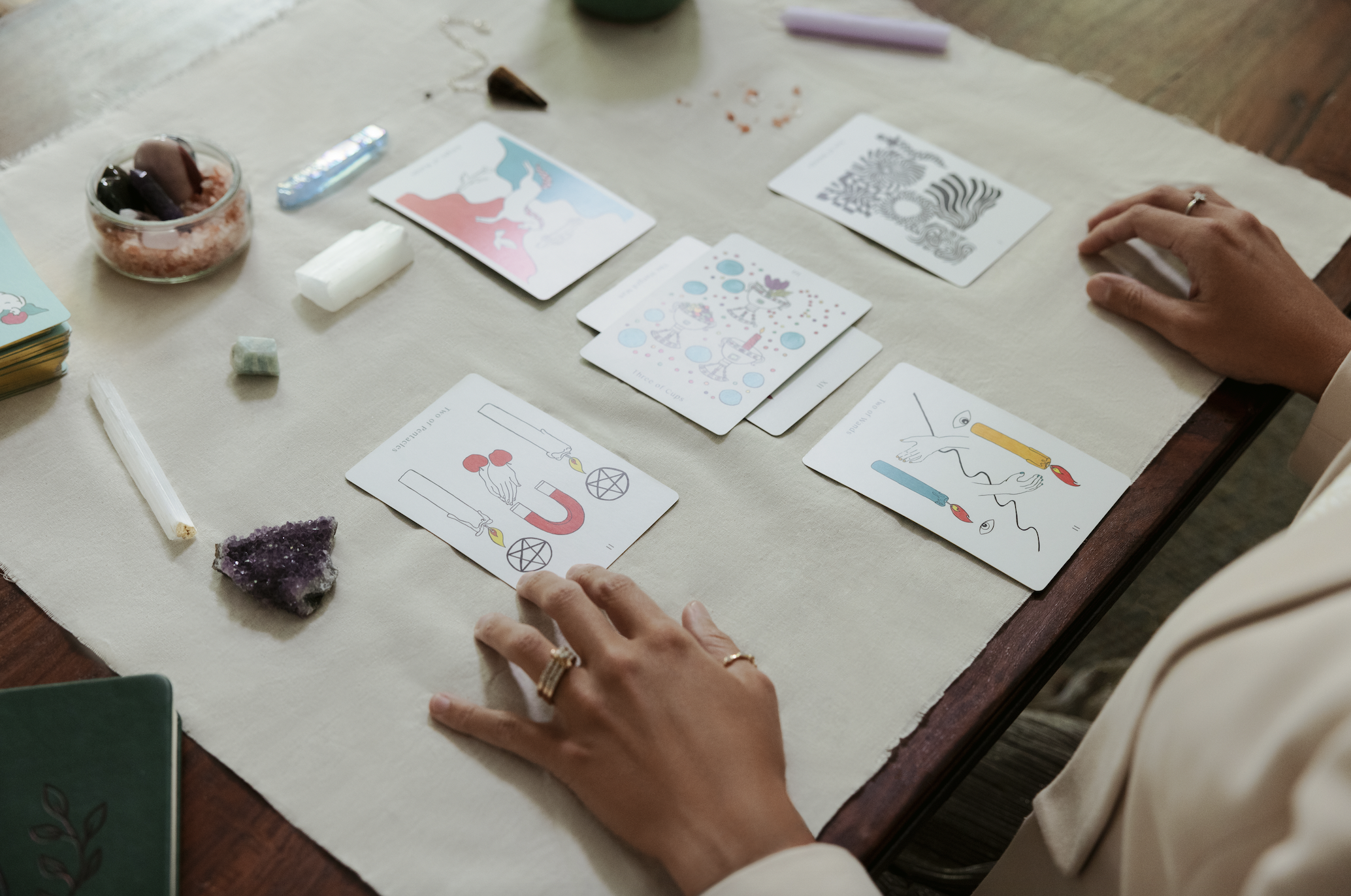 A person is sitting at a table, with hands resting in front of a spread of tarot cards laid out in front of them.