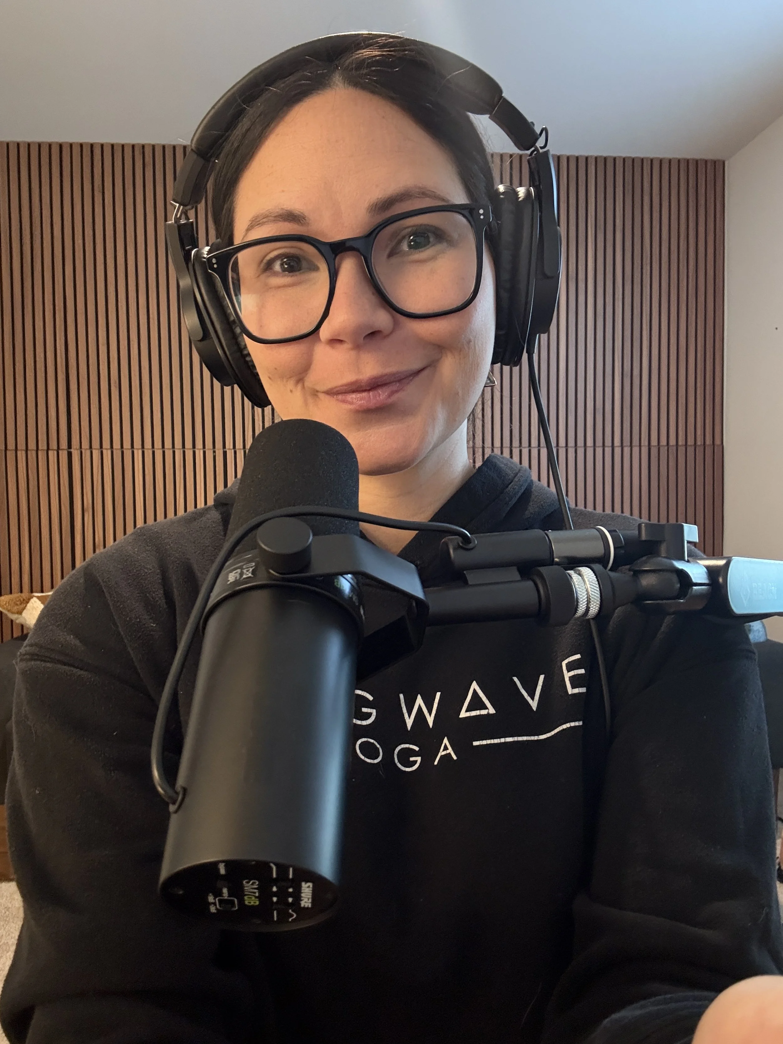 A woman with short dark hair, glasses, and headphones smiling at the camera, sitting in front of a microphone, with wooden wall paneling behind her.