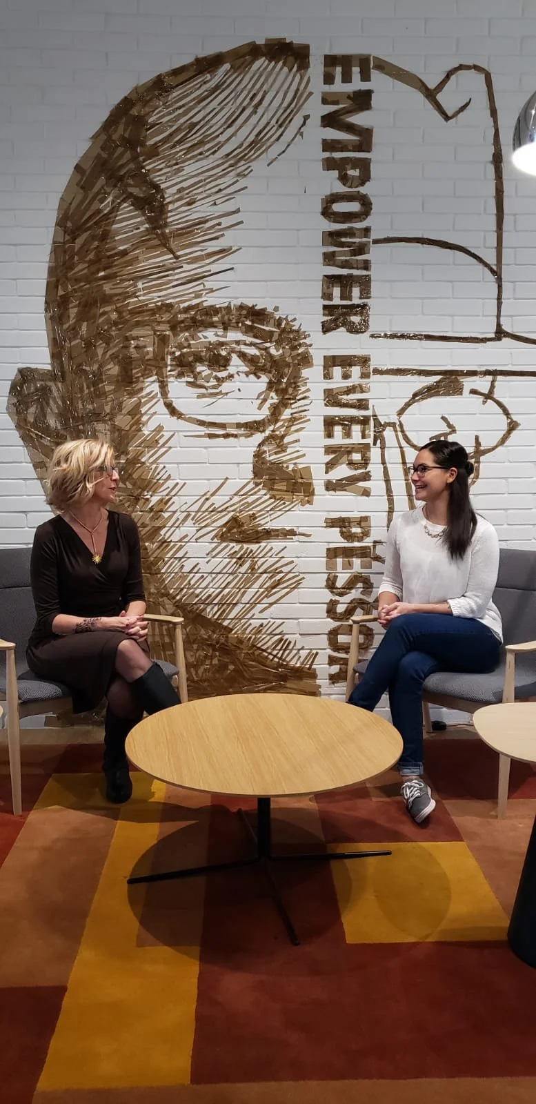 Two women sitting and talking in an office or lounge area, with a large art piece on a white brick wall behind them featuring a woman's face and the words 'Empower Every Passion'.