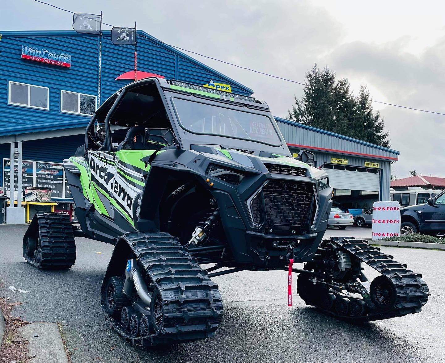 A small, tracked off-road vehicle in front of an auto repair shop with a blue exterior. The vehicle is painted black with bright green and white accents, and has the word 'Arctic' on its side. It features a boxy design, large treads for tracks, and an industrial front with a visible turbo engine.
