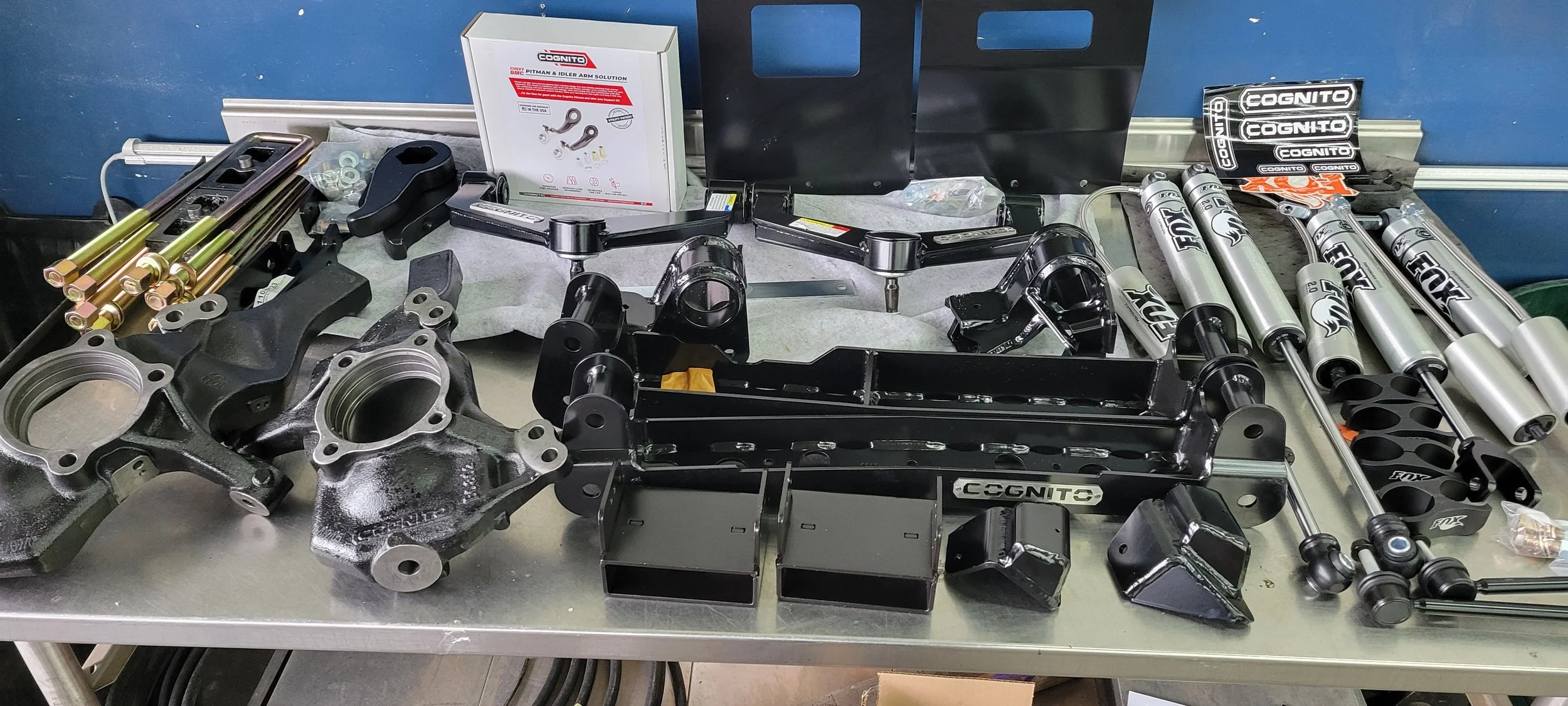 A workbench with motorcycle parts including shock absorbers, engine components, brackets, and various tools, along with a box of arm solutions, set against a blue wall.