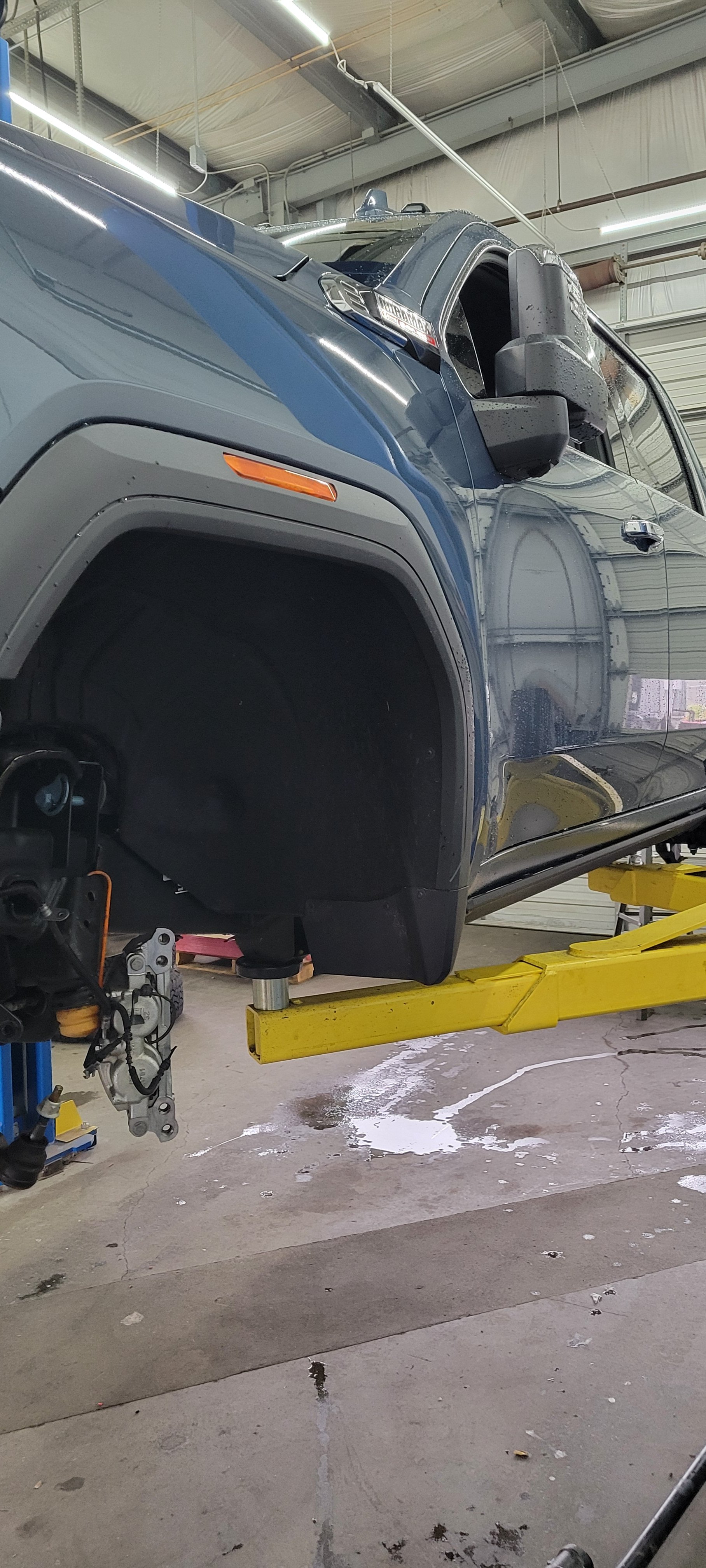 A blue SUV is lifted on a hydraulic lift inside a garage or workshop, with one wheel removed and the vehicle’s front left corner exposed.