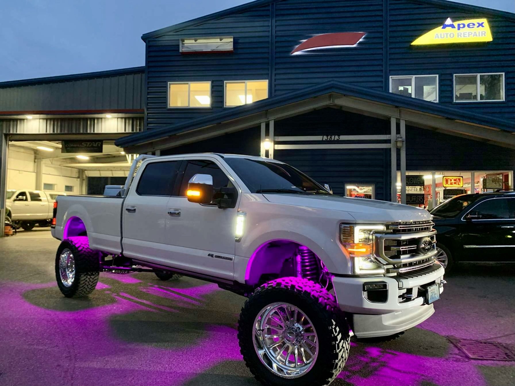 White lifted pickup truck with purple underglow lights parked in front of auto repair shop at dusk. Building has blue siding, illuminated windows, signs for auto repair, and an open sign in the window.