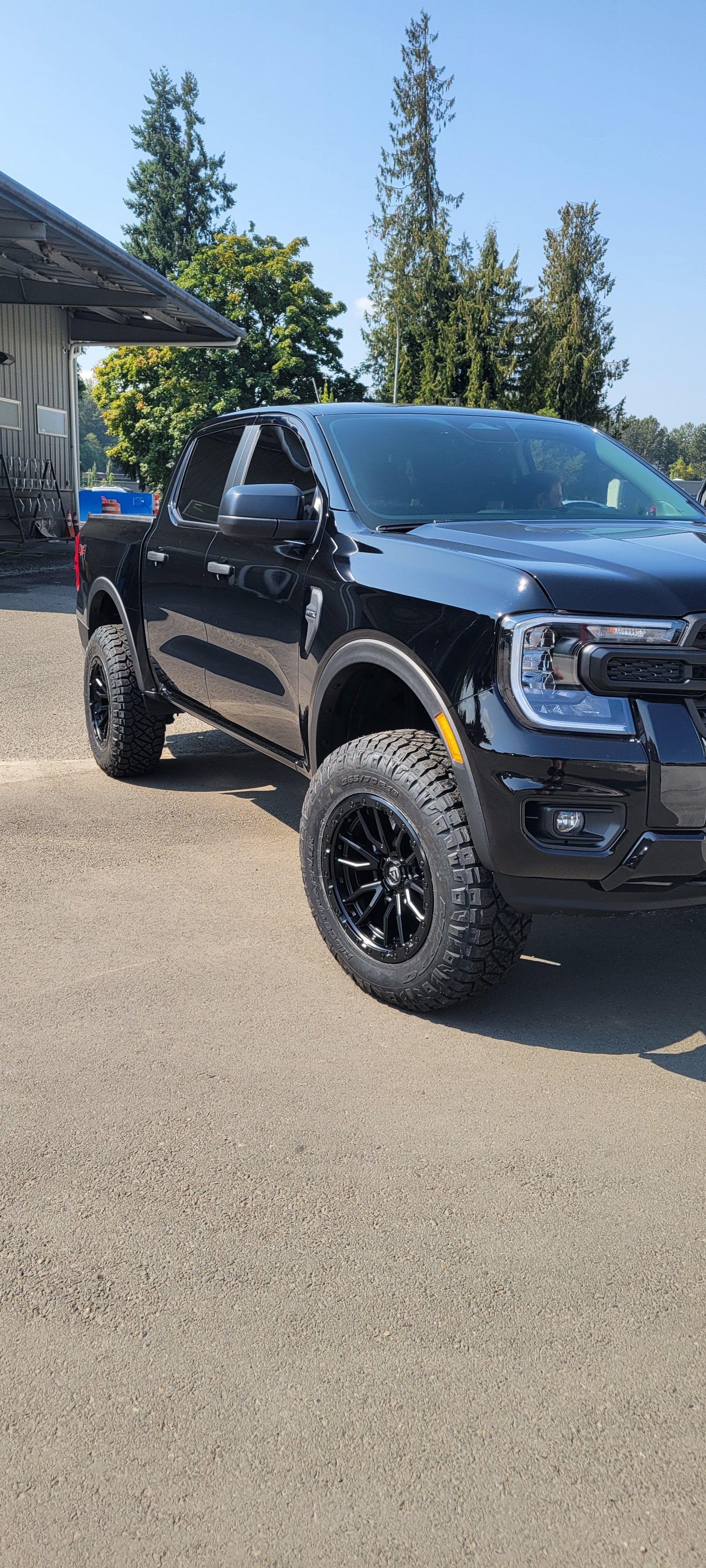 Black pickup truck with off-road tires parked on a paved surface under a clear blue sky, with trees and a building in the background.