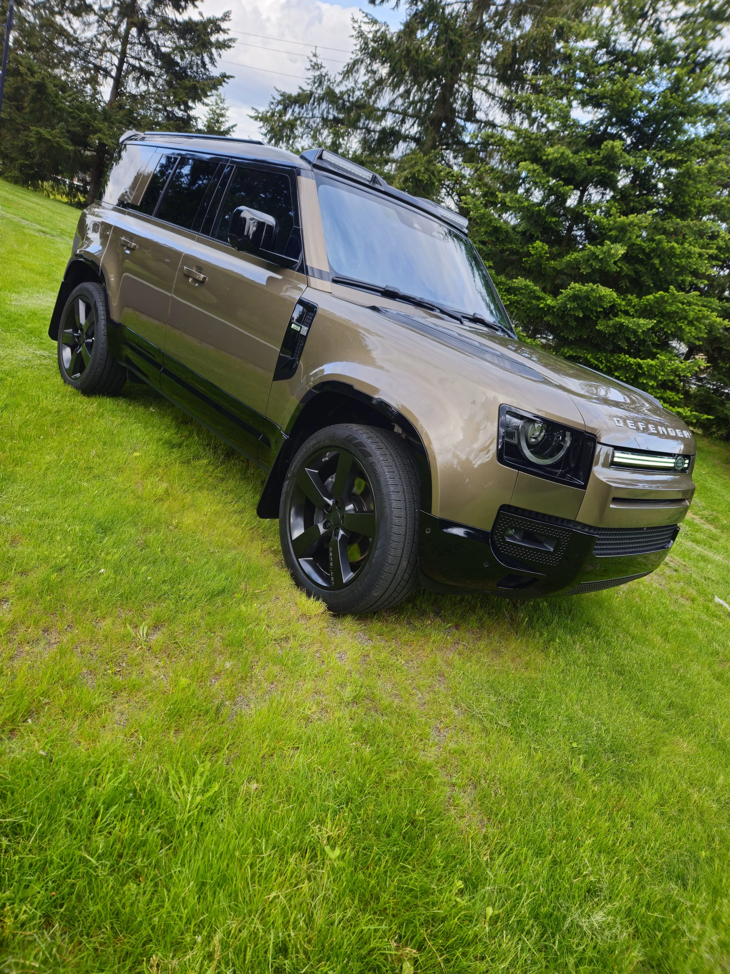 A tan Land Rover Defender SUV parked on a grassy field with tall trees in the background.