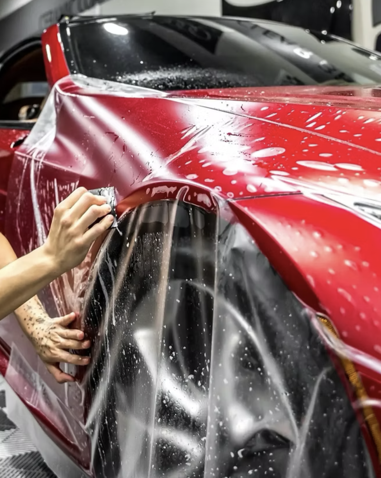 A person applying protective film or wrap to a red and black sports car in a garage or workshop.