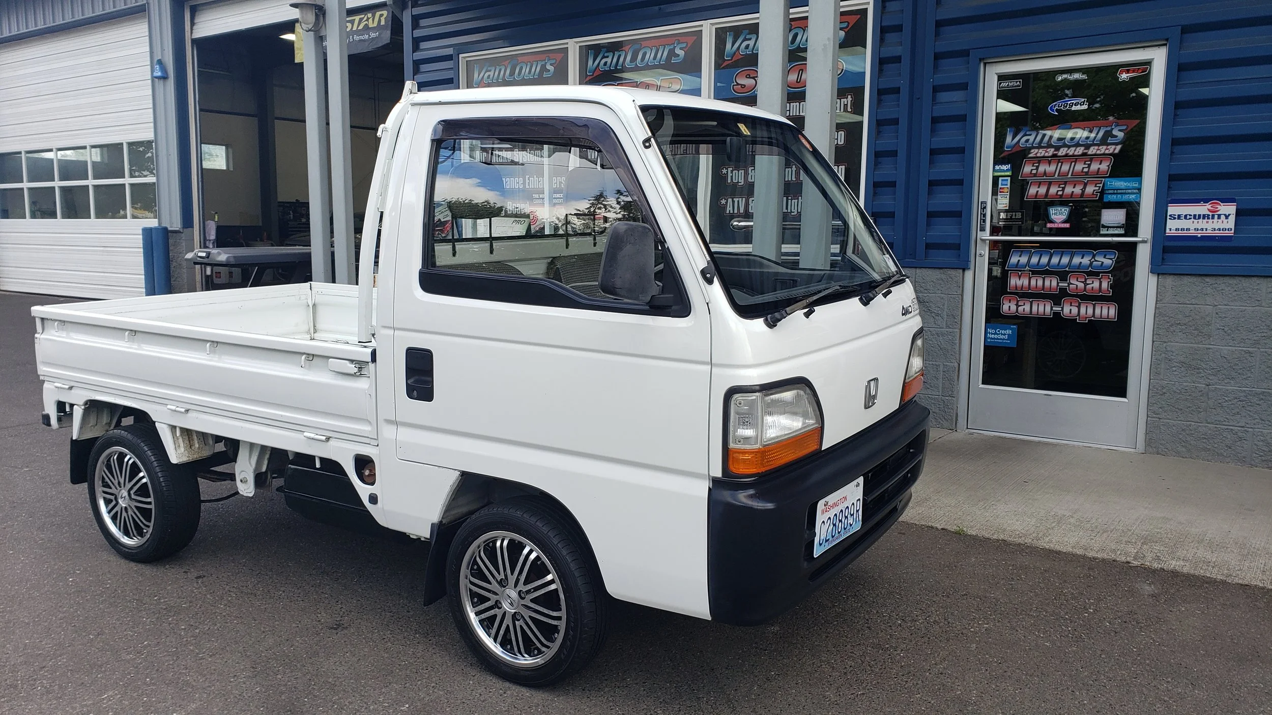 White mini truck parked outside an automotive shop with blue siding and glass windows and door, signage on the windows reading VanTour's. The shop offers services like fog lights, ATVs, and LED lighting. The truck has a Washington license plate and c