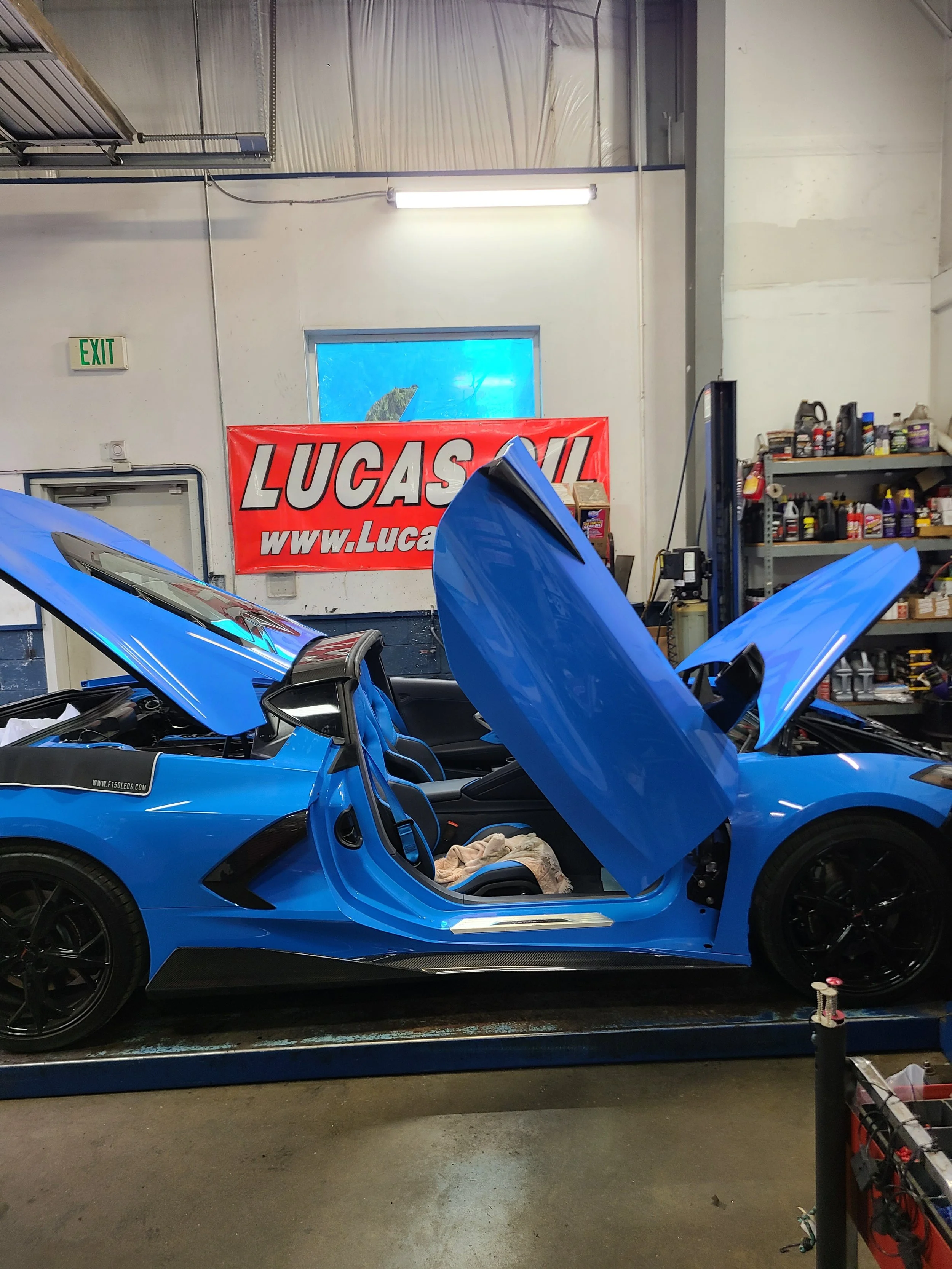 A bright blue sports car with its doors open in a garage, with tools and supplies on shelves in the background.