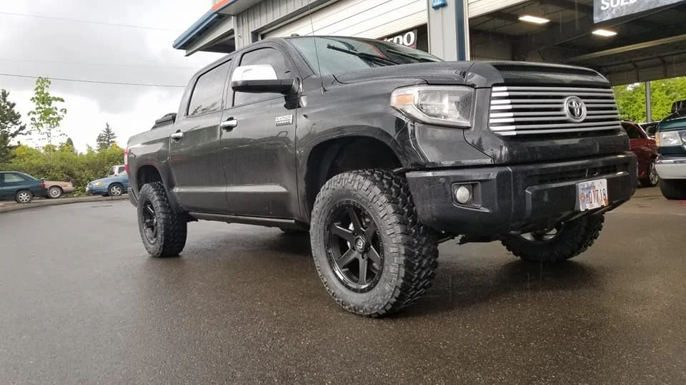A black Toyota Tundra pickup truck with large off-road tires and black rims parked in a dealership lot.