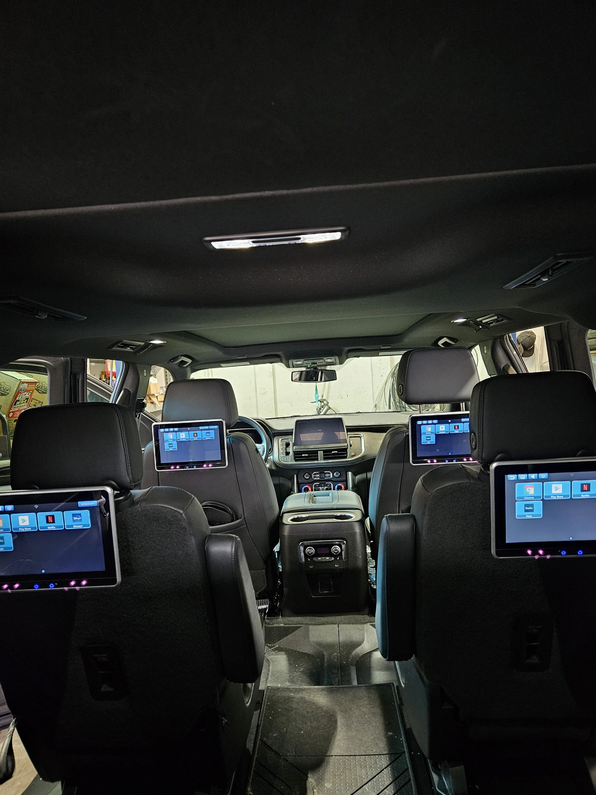 Interior of a vehicle with black seats and multiple screens mounted on the back of the headrests, showing an app grid, with a steering wheel and dashboard visible in the front.