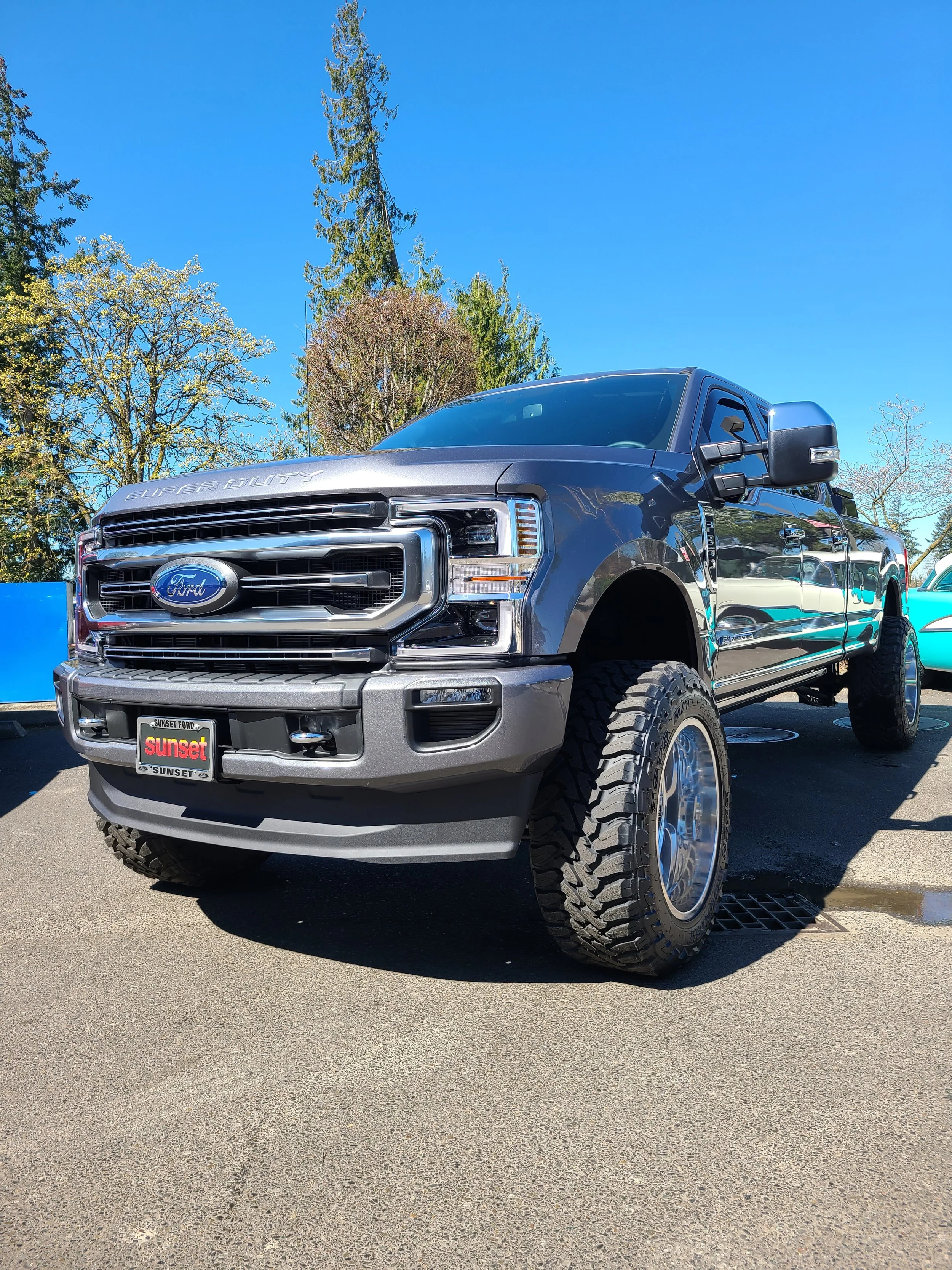 A black Ford Super Duty pickup truck with large tires parked on a paved lot under a clear blue sky. There are trees in the background and a blue structure to the left.