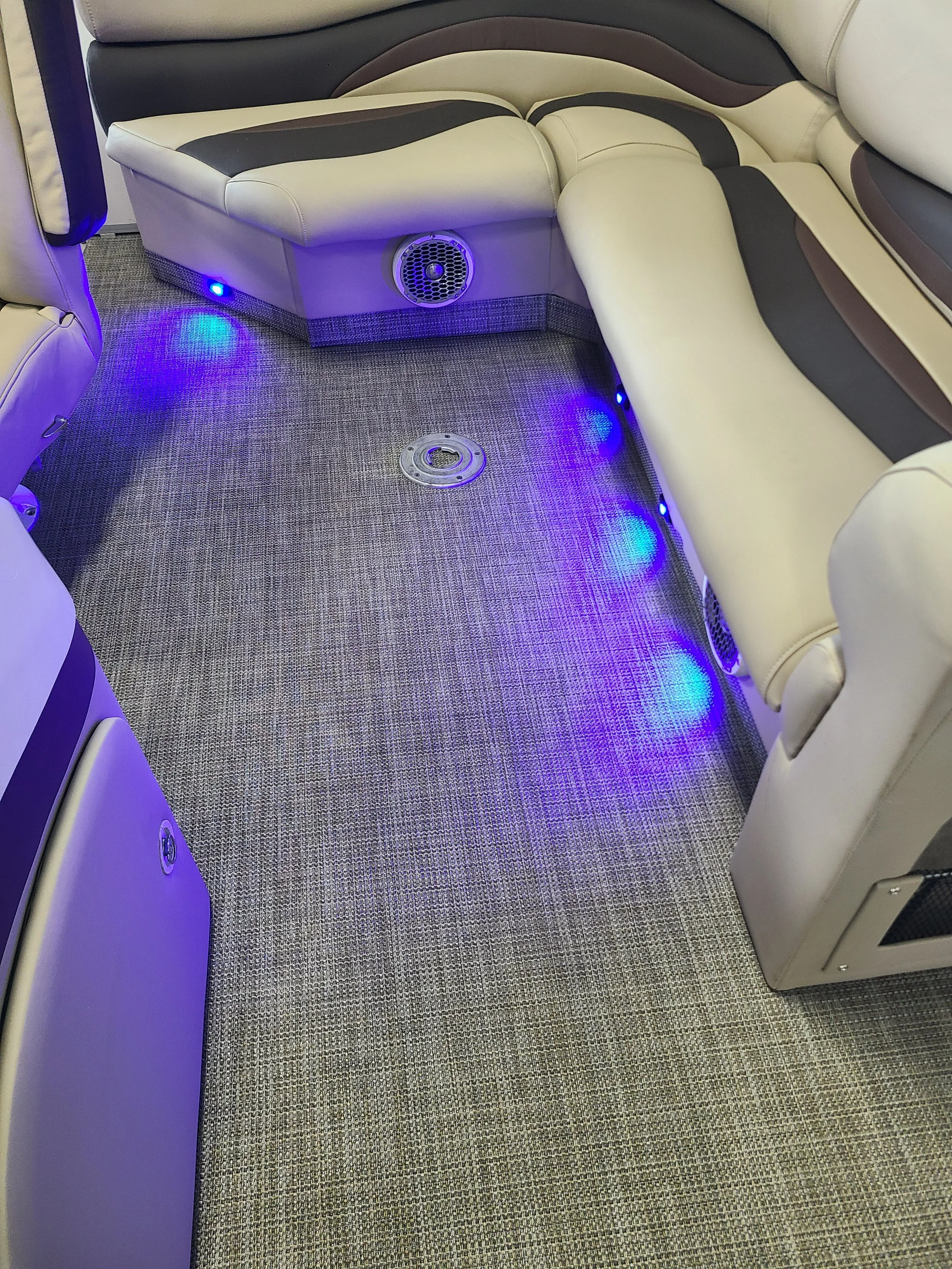 Interior of an airplane with beige leather seats, a textured gray carpet, and blue LED lighting along the floor. There is a gray metallic vent cover on the floor and a small speaker or vent on the side of the seat.