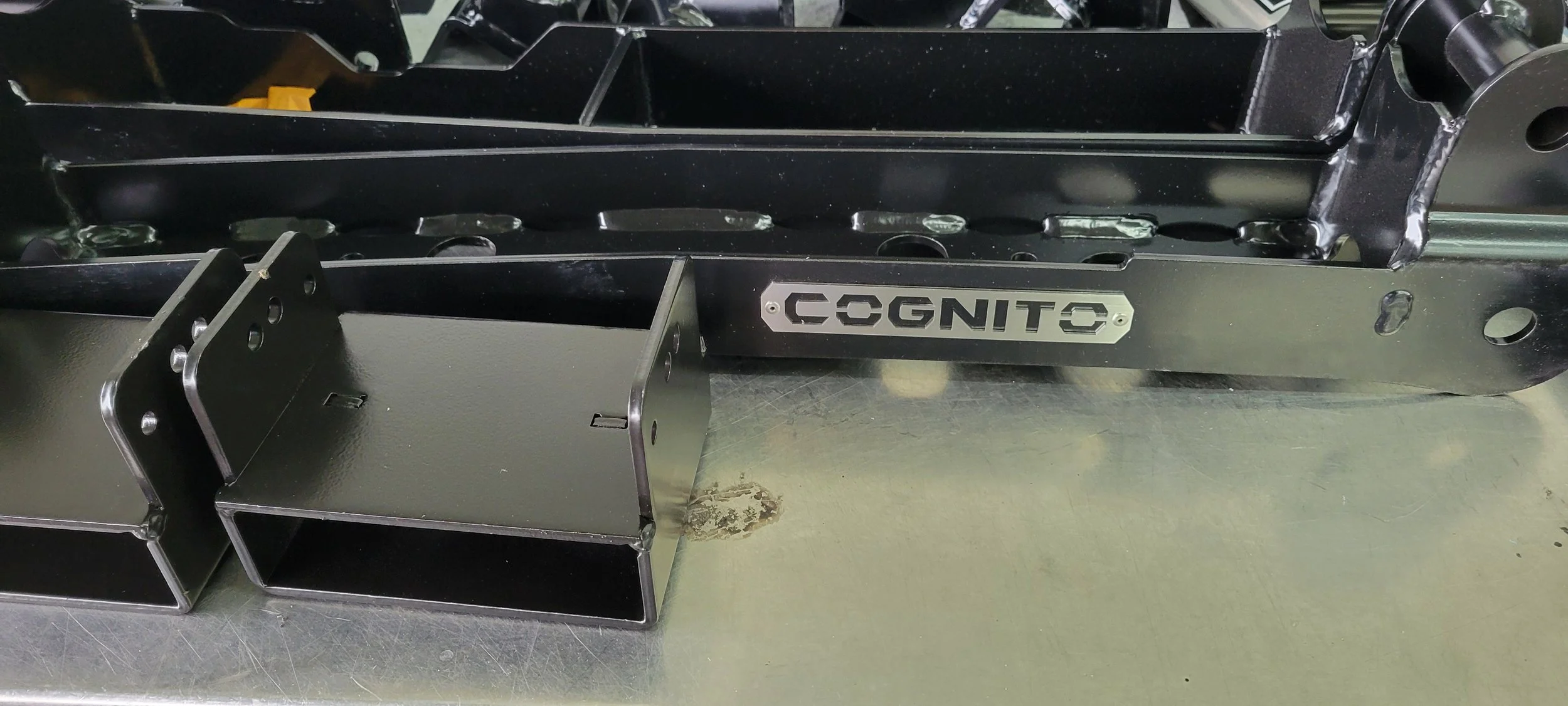 Close-up of a black metal frame with the label 'COGNITO' attached to it, featuring multiple holes and structural components; possibly part of a piece of machinery or equipment.