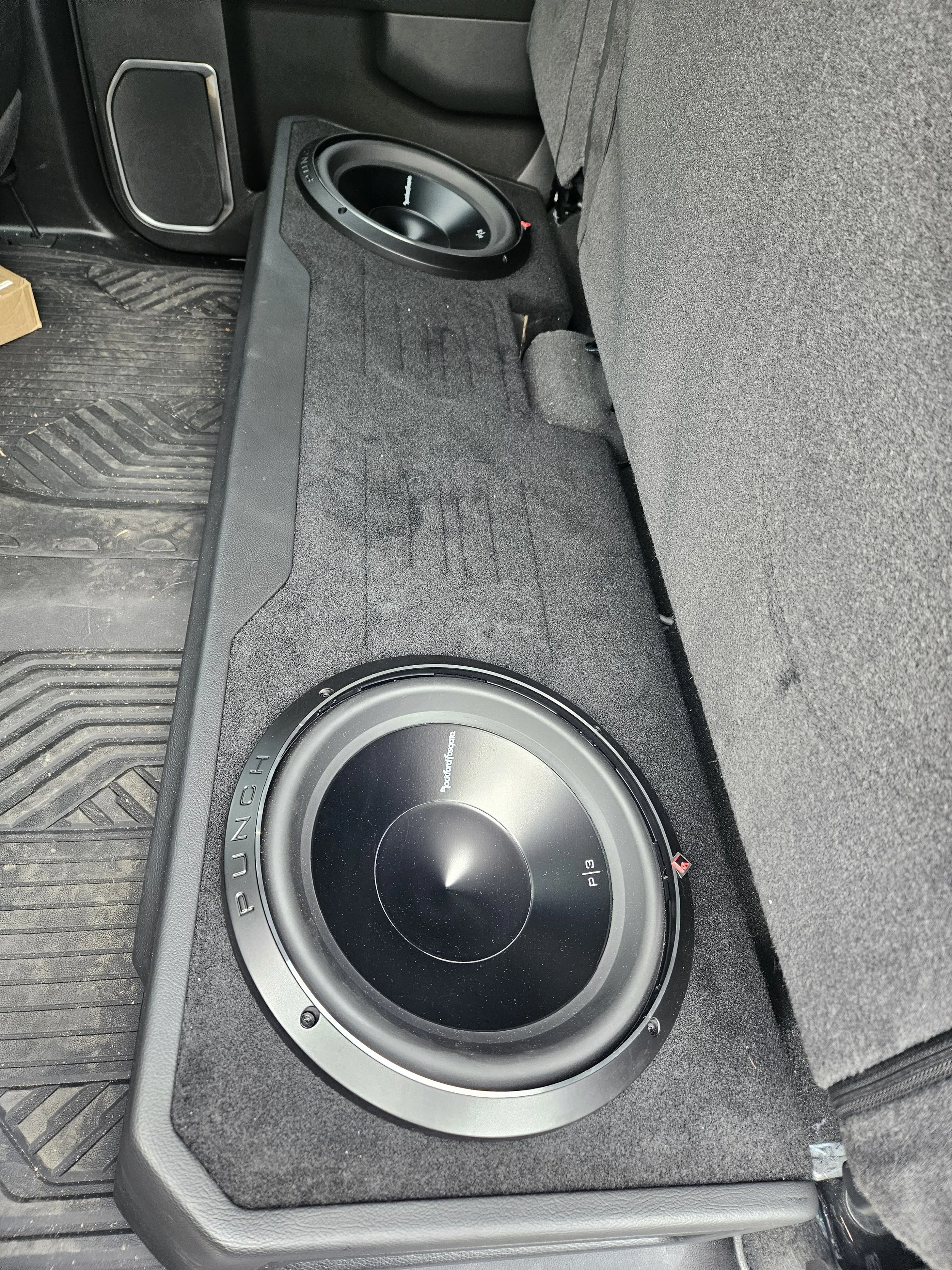 A close-up view of a car's interior showing a large subwoofer speaker installed in the trunk area, with carpeted surface around it.