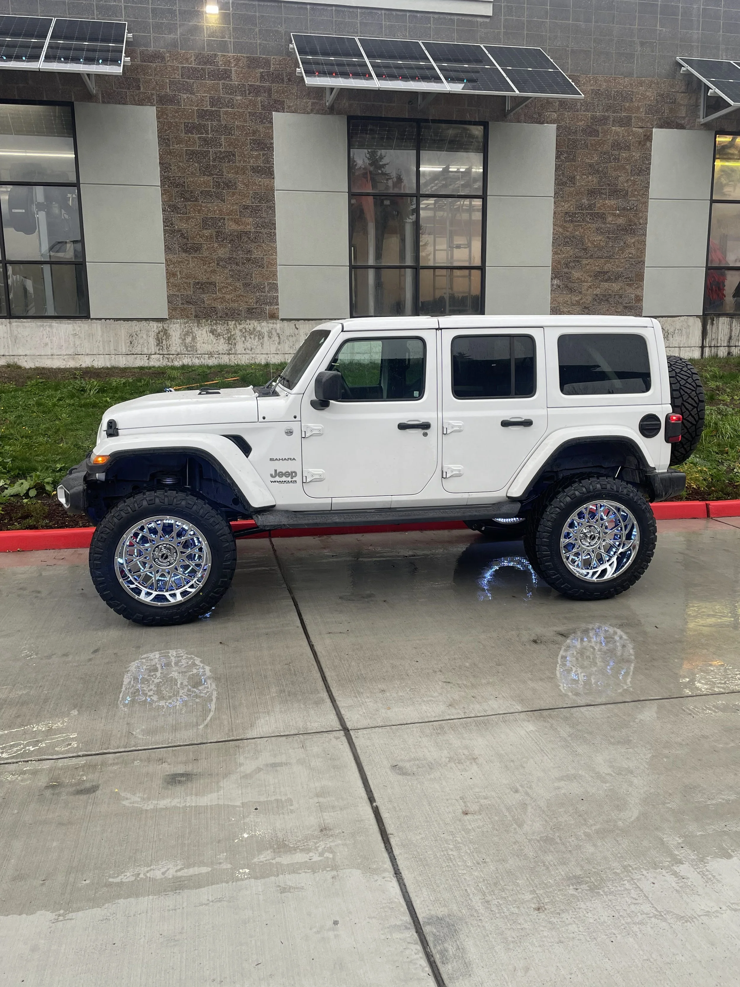 A white Jeep Wrangler Sahara parked on wet pavement next to a curb with red paint and dirt, with a building featuring solar panels and large windows in the background.