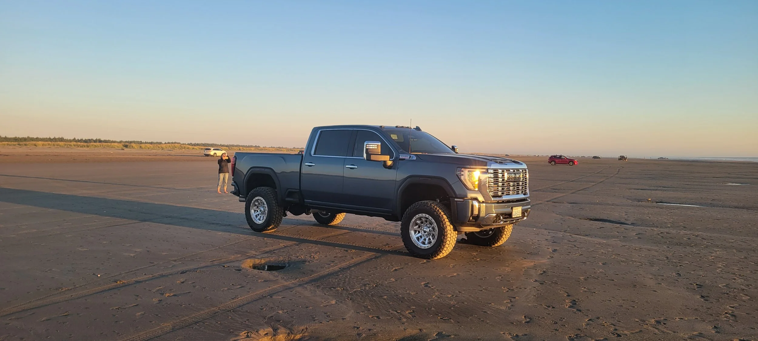 A large black pickup truck parked on a flat, sandy area at sunset, with a person standing nearby and several other vehicles in the distance.