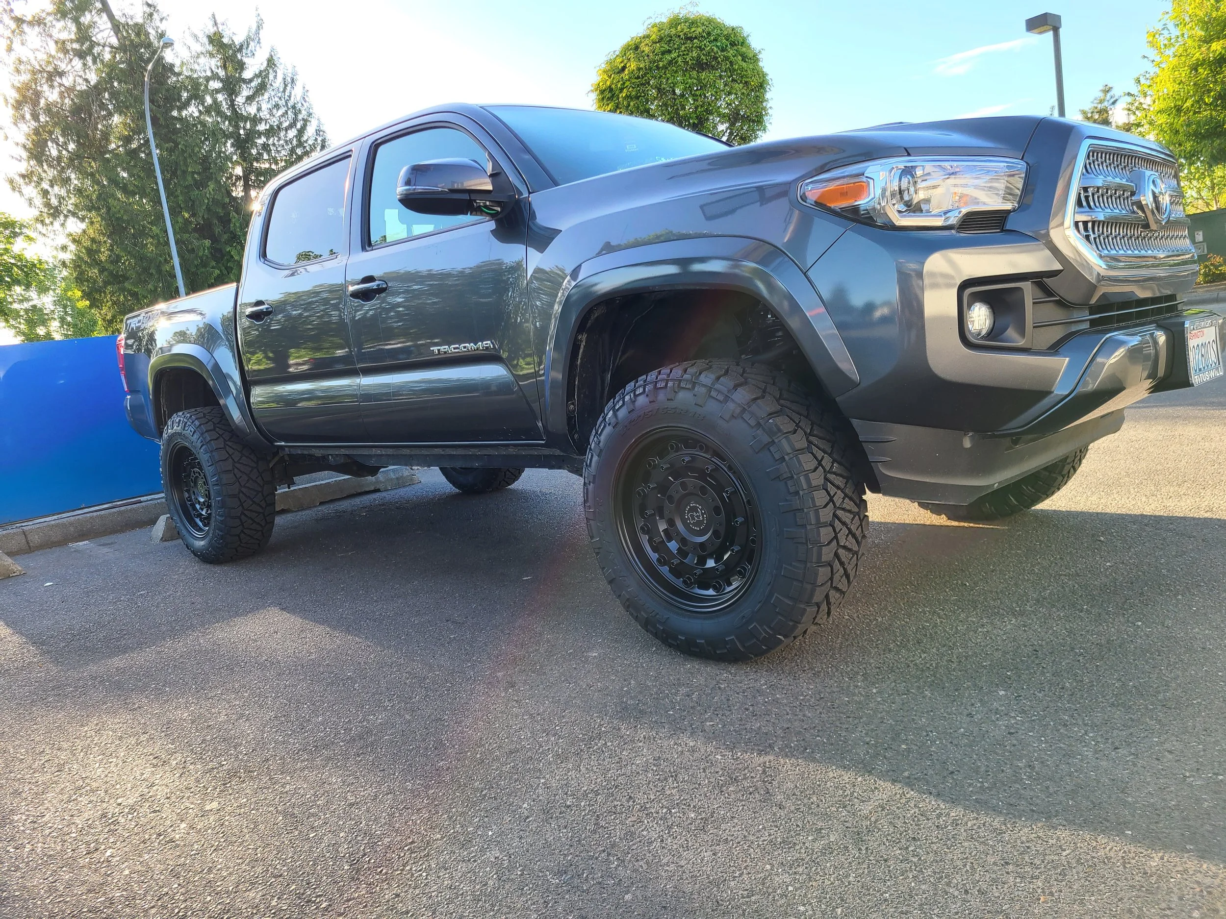 A black Toyota Tacoma pickup truck parked in a parking lot with trees and a blue fence in the background.