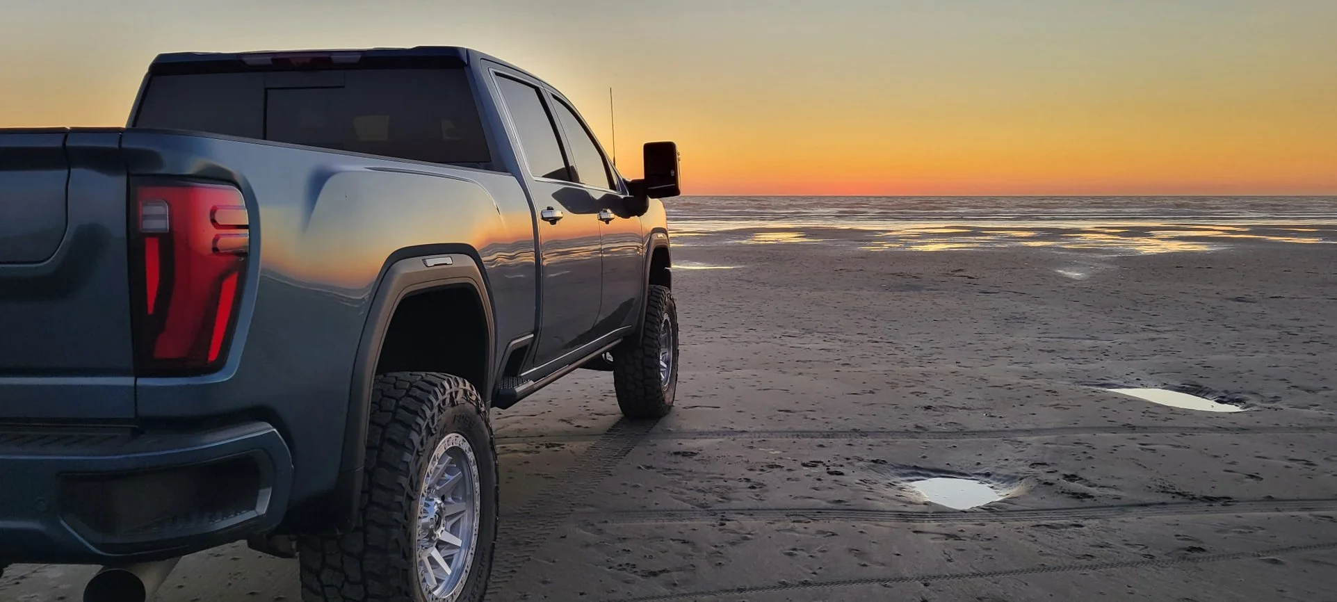 A blue pickup truck parked on a sandy beach at sunset, with calm ocean waves in the background and small pools of water on the sand.