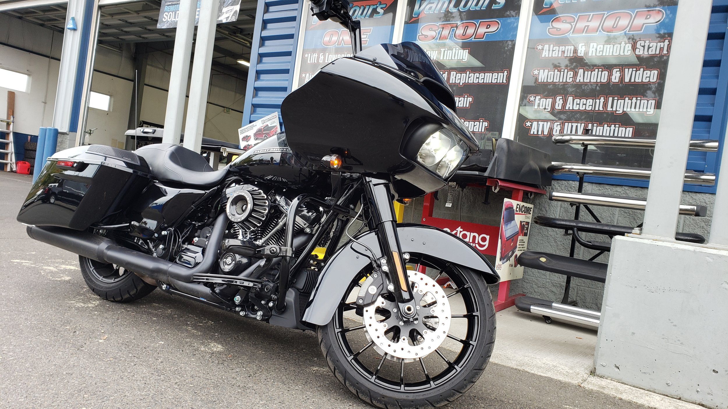 A black and white touring motorcycle parked outside an auto repair shop, with signs advertising various car services in the background.