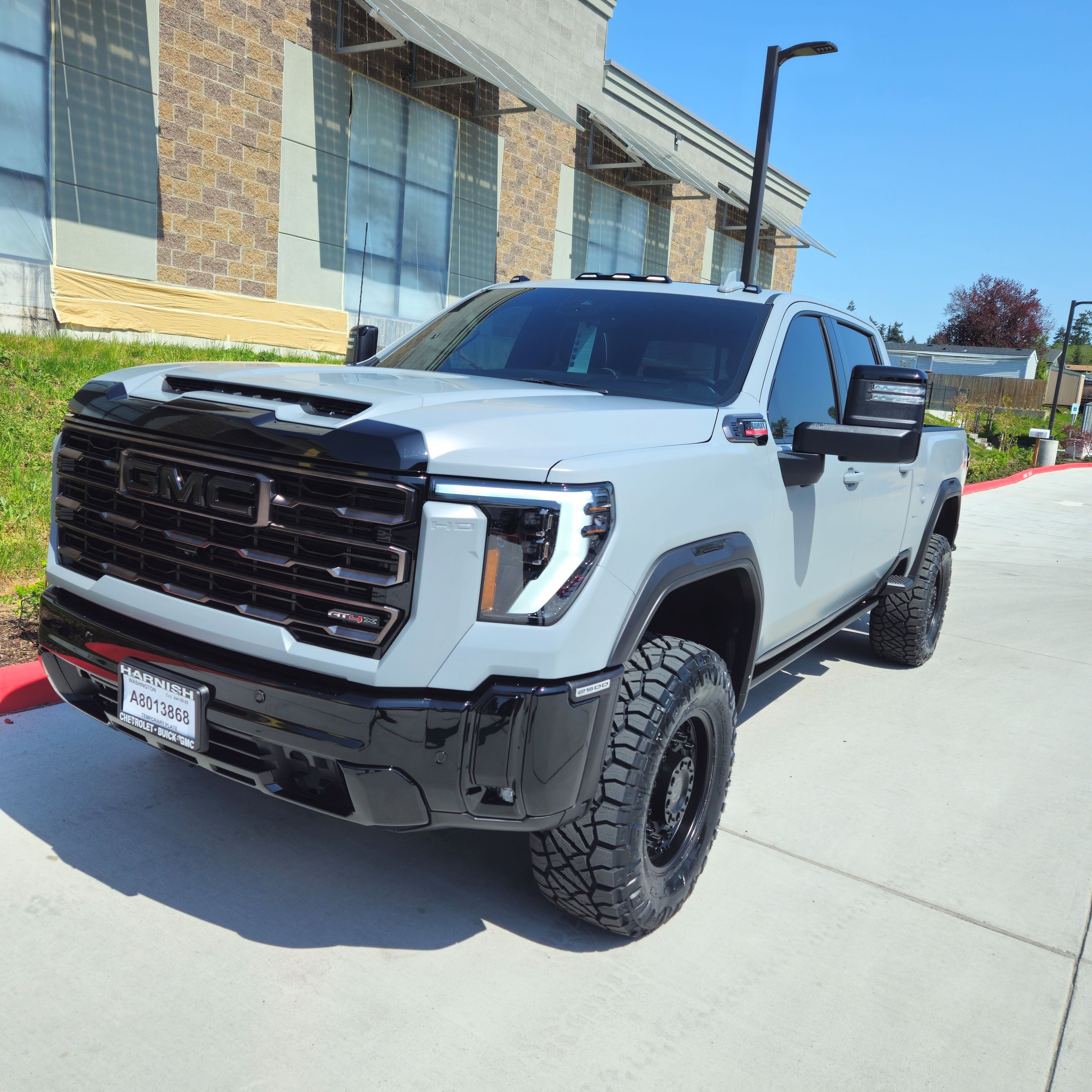 A white GMC truck with large off-road tires parked outside a building with brick and glass walls under a clear blue sky.