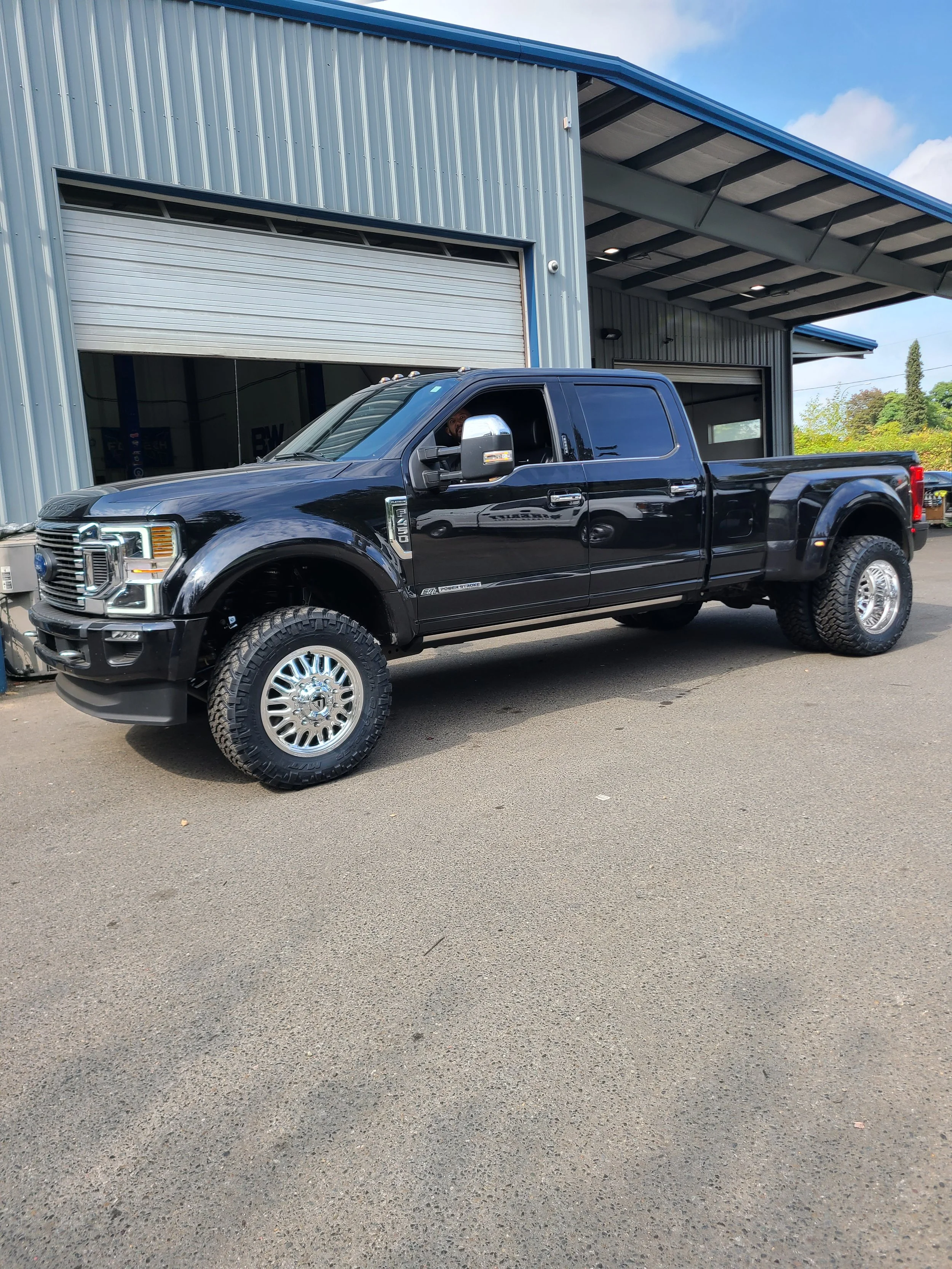 A black Ford F-250 pickup truck parked in front of a gray industrial building with a blue roof, partially open garage door, and a man sitting inside.