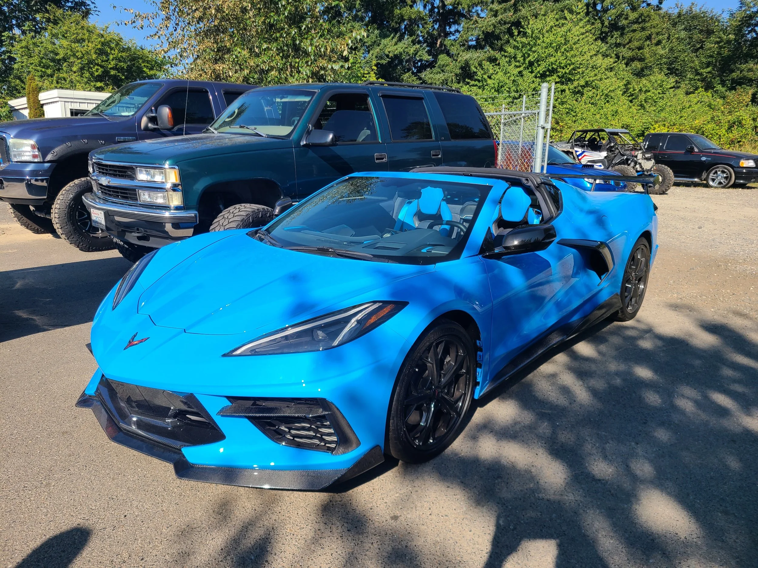 A bright blue sports car in the foreground with a sleek design, side mirrors, and a low profile. Behind it, there are several other vehicles including a dark green SUV, a black sedan, and another black vehicle, parked outdoors in a lot with a chain-l