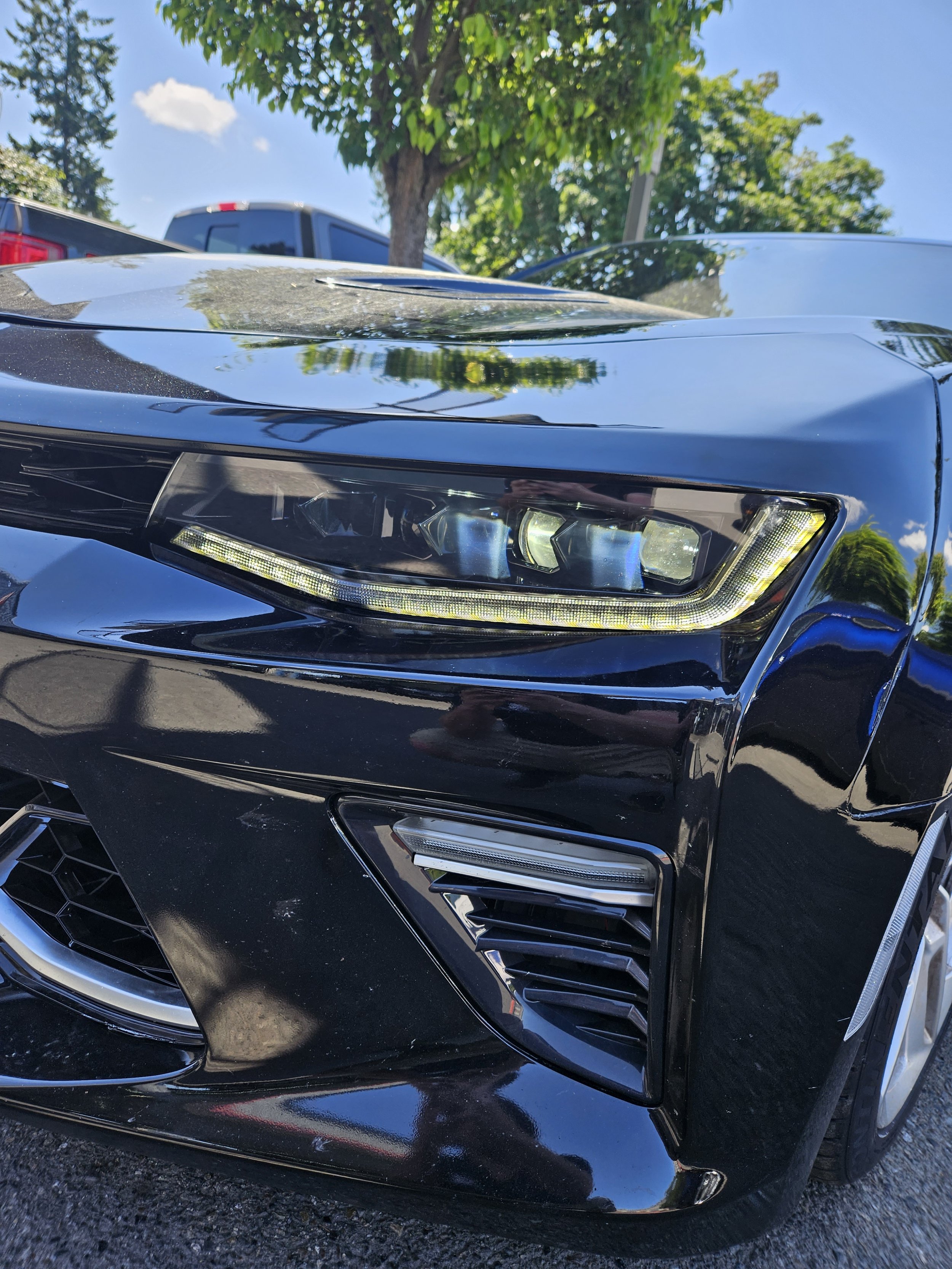 Close-up of the front of a shiny black car, focusing on the headlight, grille, and front bumper, with trees and a blue sky in the background.