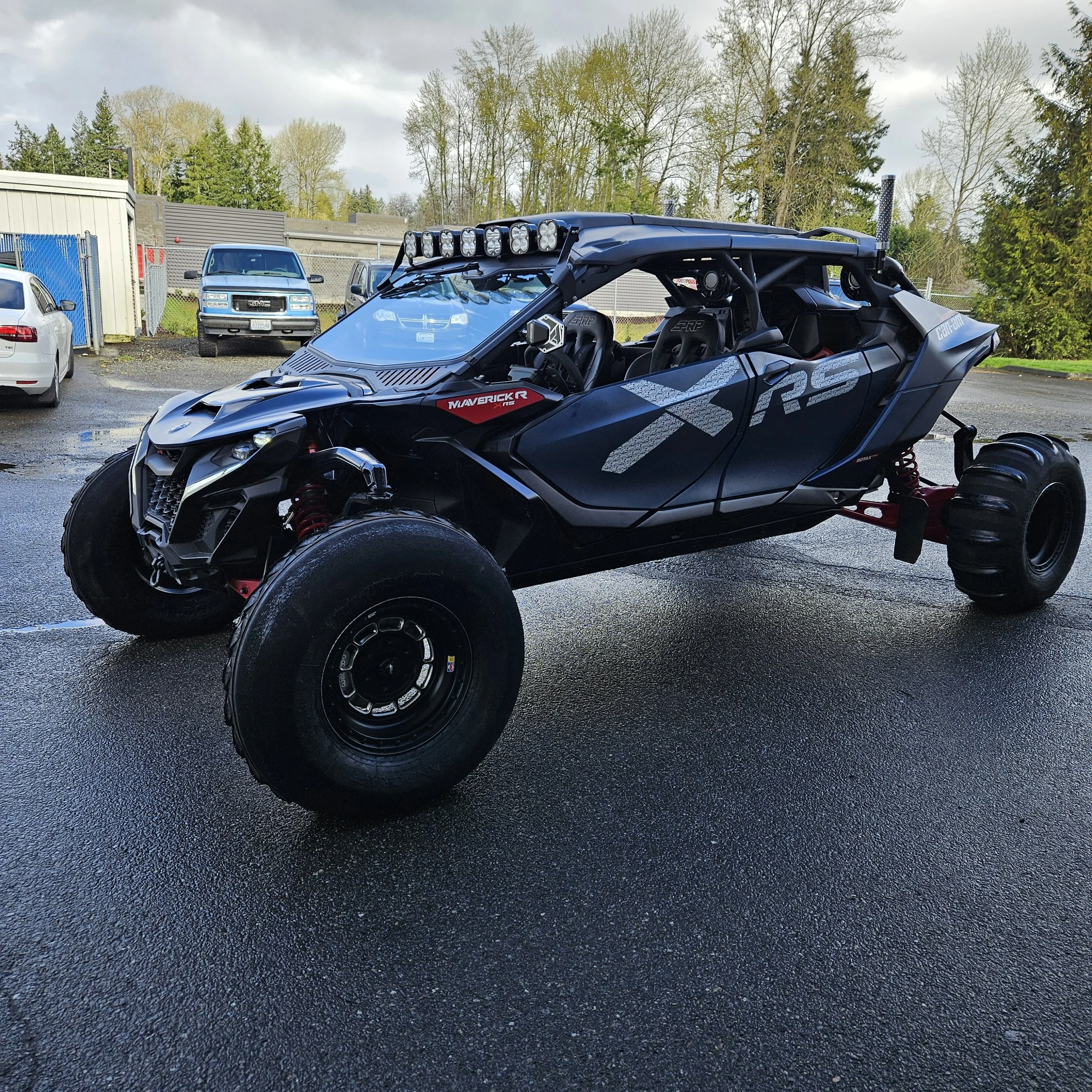 Off-road vehicle with multiple bright lights mounted on top, parked outdoors on a wet surface, with trees and cloudy sky in the background.