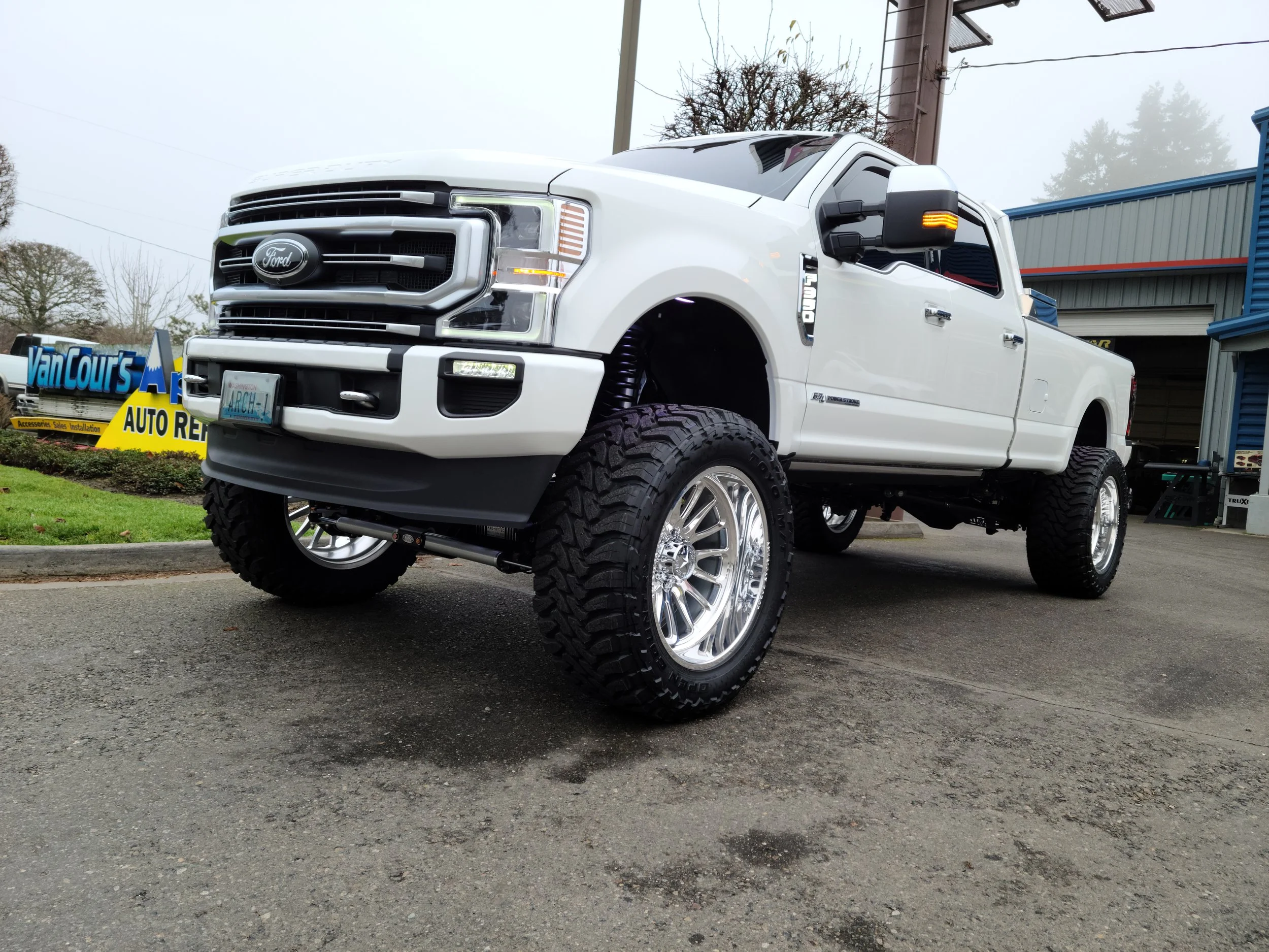 A white Ford pickup truck parked outdoors on a cloudy day, with large off-road tires, custom wheels, and a lift.