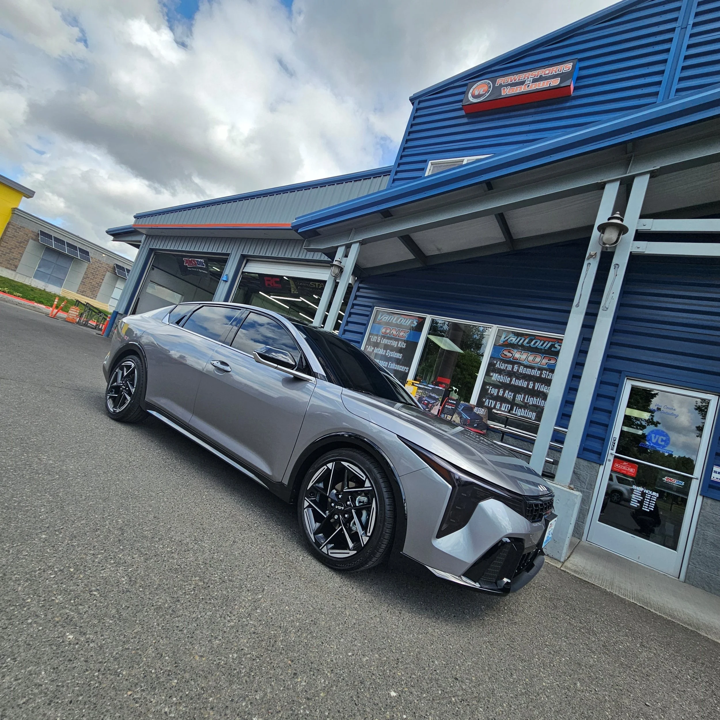 A silver car parked in front of a blue building with signage for Powersports by VanCourts, specializing in lift and lowering kits, air intake systems, and other automotive performance parts. The building has a metal exterior with a sloped roof, and the parking lot has some orange safety cones and a neighboring yellow building.