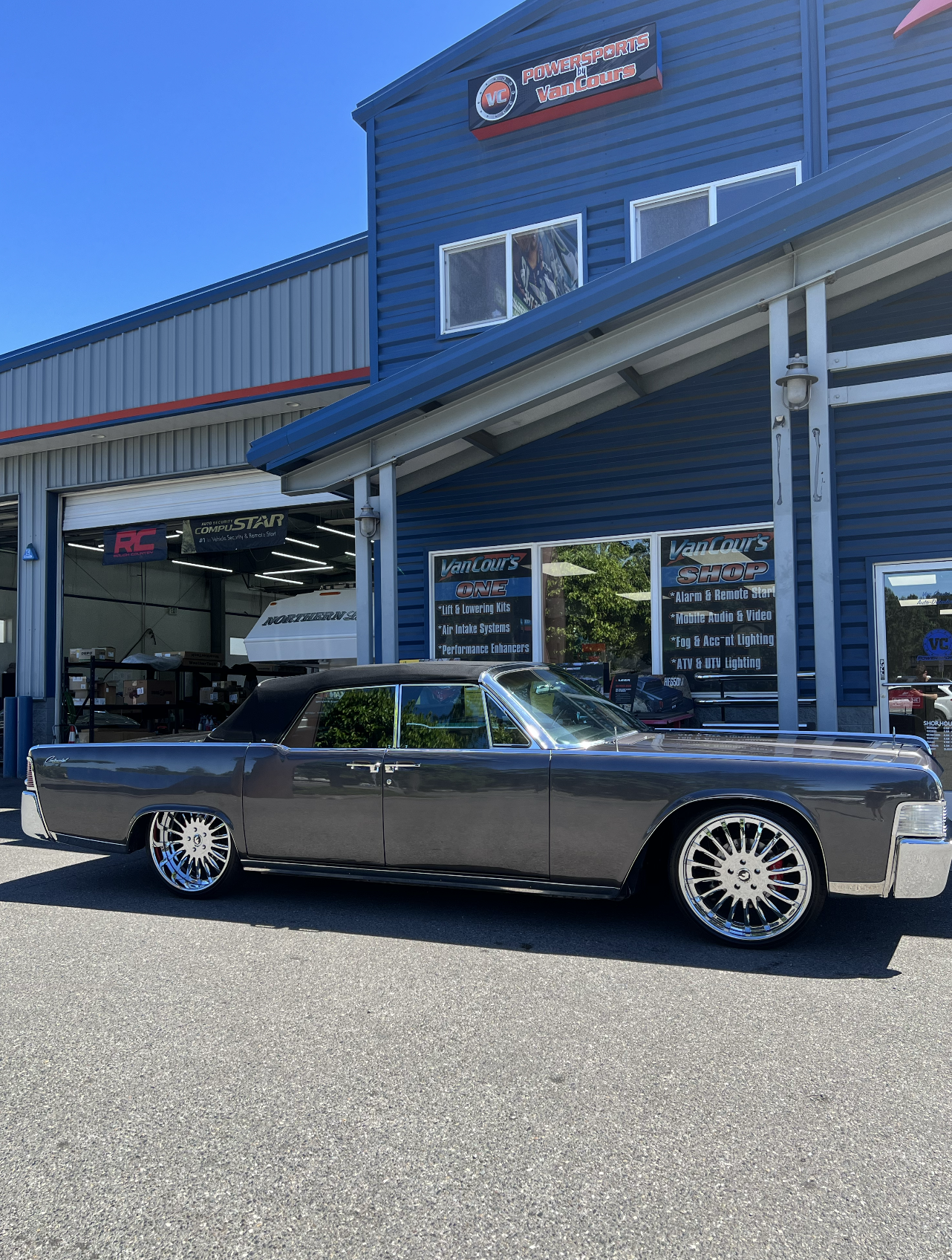 A classic black convertible car with large, shiny alloy wheels parked outside an automotive store named Van Cour's Powersports, which sells vehicle accessories and performance parts, with a bright blue sky overhead.