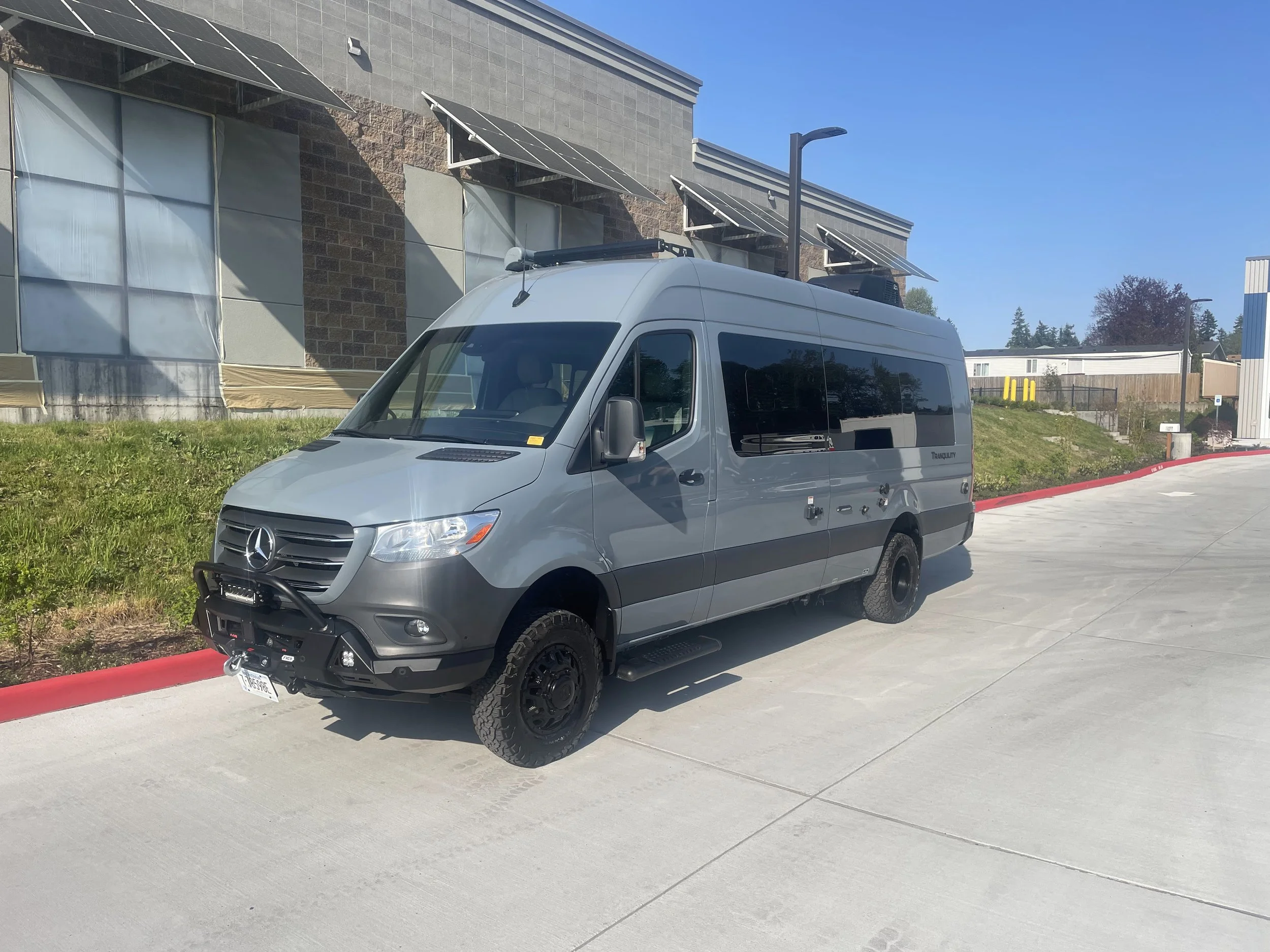 A silver Mercedes-Benz van parked on a concrete sidewalk in front of a modern building with solar panels on the roof.