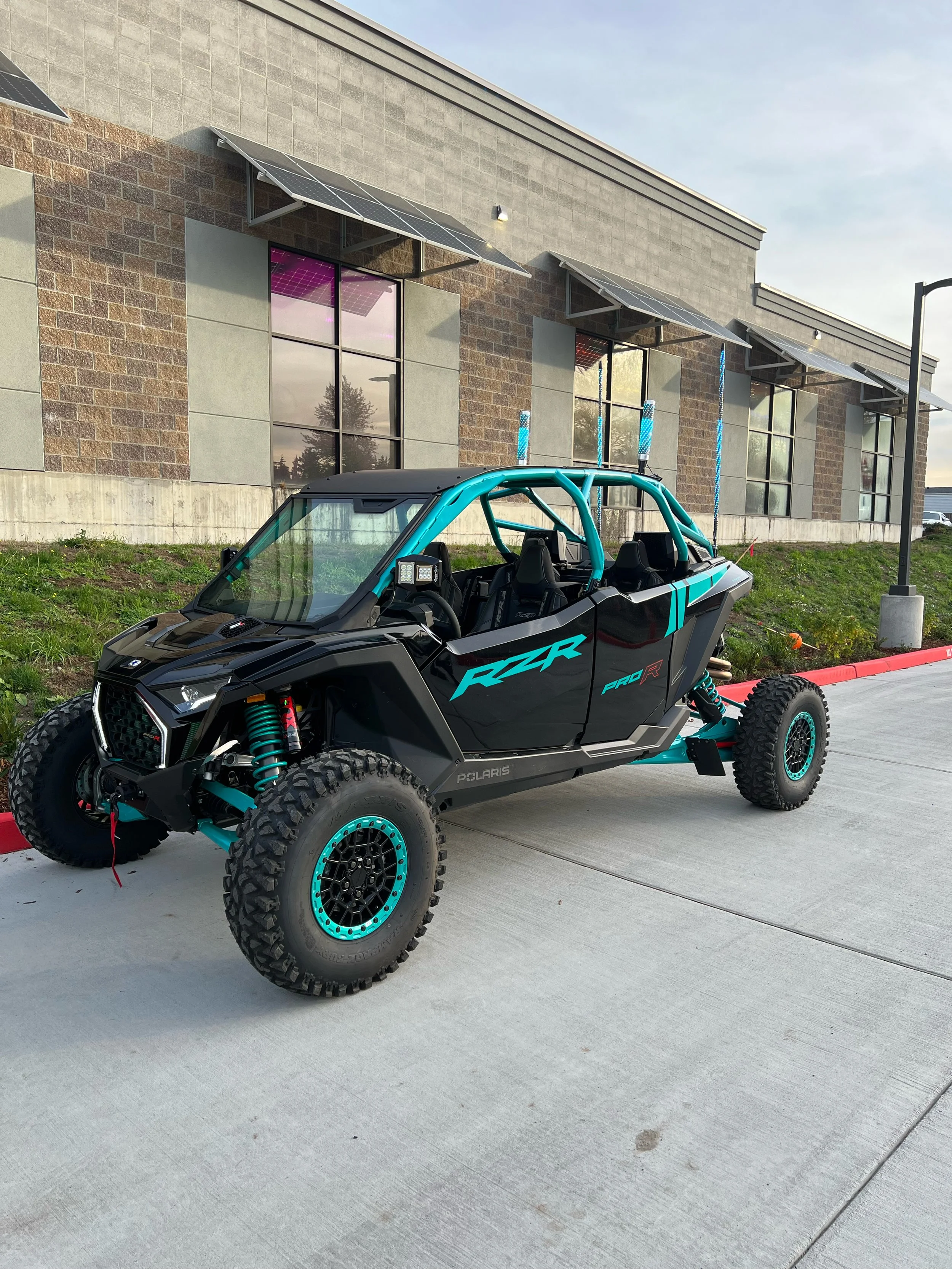 A Polaris RZR off-road vehicle with black and teal color scheme parked on a sidewalk in front of a building with large windows and solar panels on the roof.