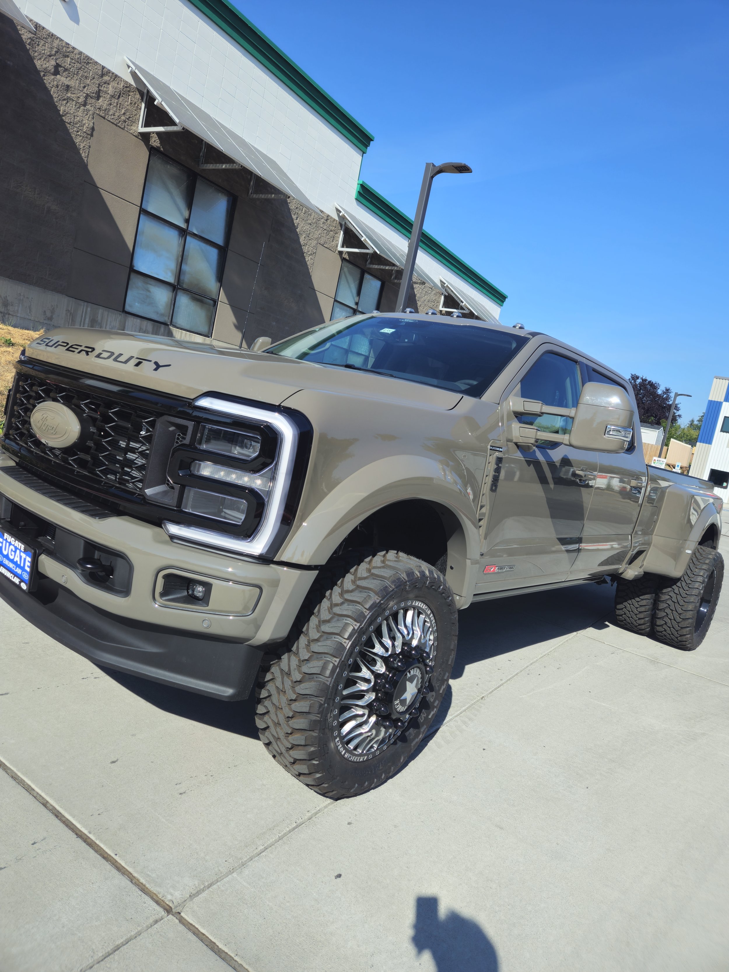 A beige Ford Super Duty pickup truck with large tires parked on a concrete sidewalk outside a commercial building. The building has a grey and white exterior with green trim and large windows. A streetlamp is visible in the background, and the sky is clear and blue.