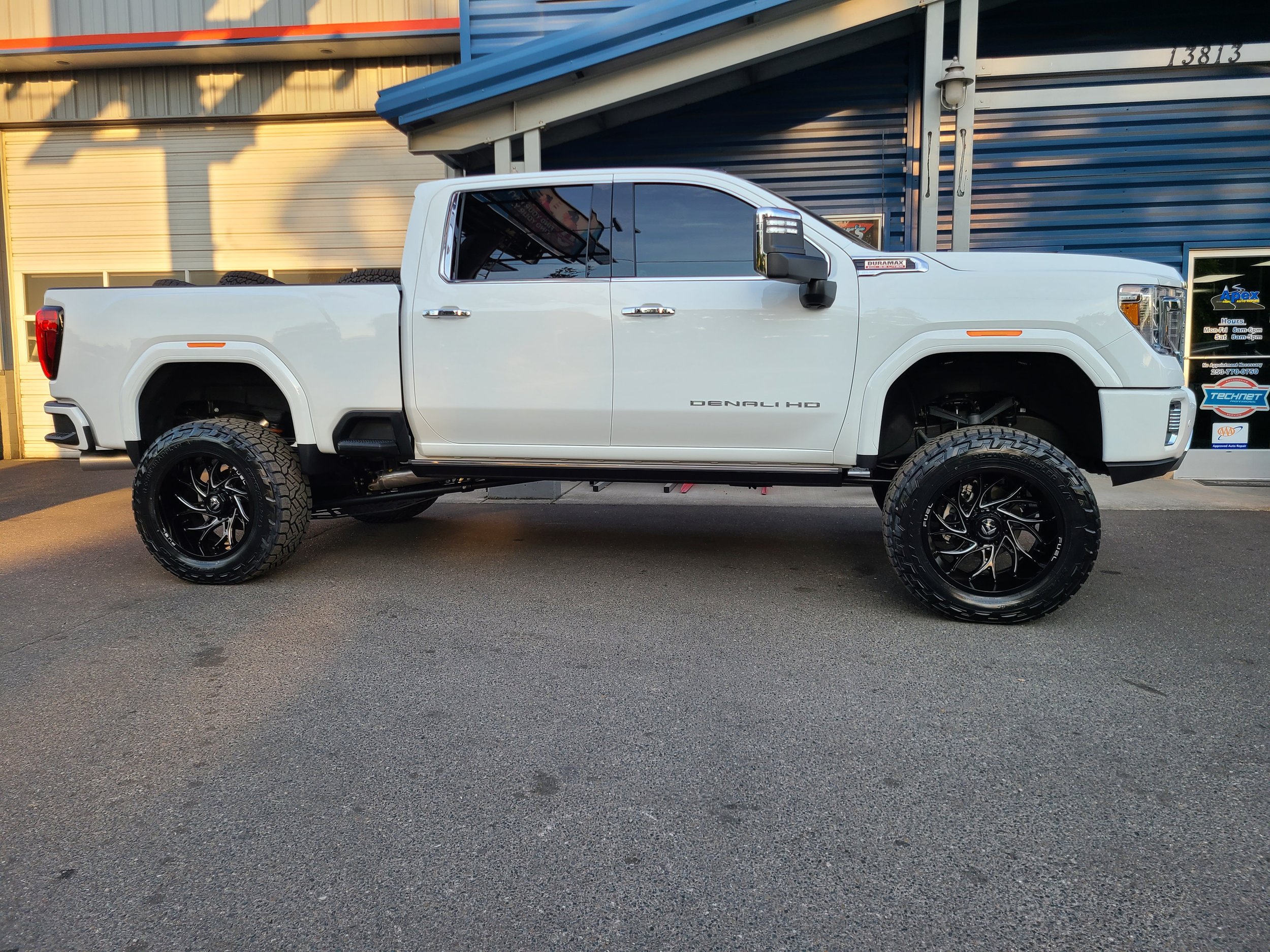 A white lifted pickup truck with large wheels parked in front of a commercial building with a blue and beige exterior. The truck has tinted windows and visible tires in the truck bed.