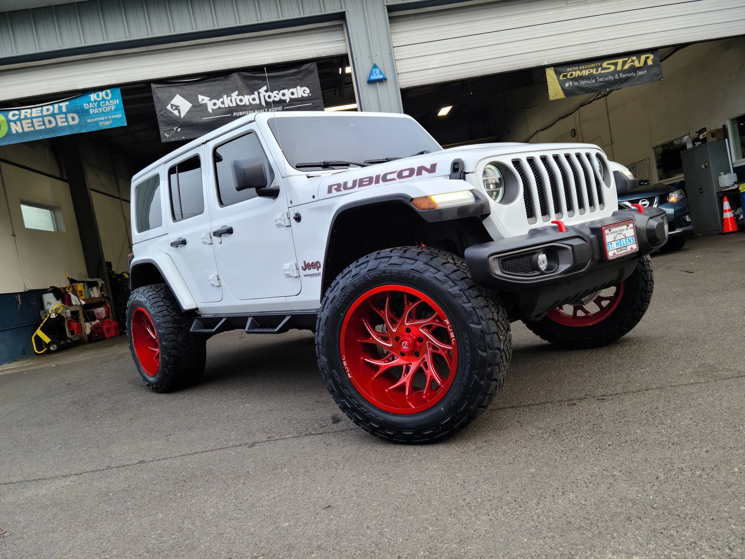 A white Jeep Rubicon SUV with red rims parked inside a garage next to other vehicles and tools, with banners hanging from the ceiling advertising car security and audio brands.