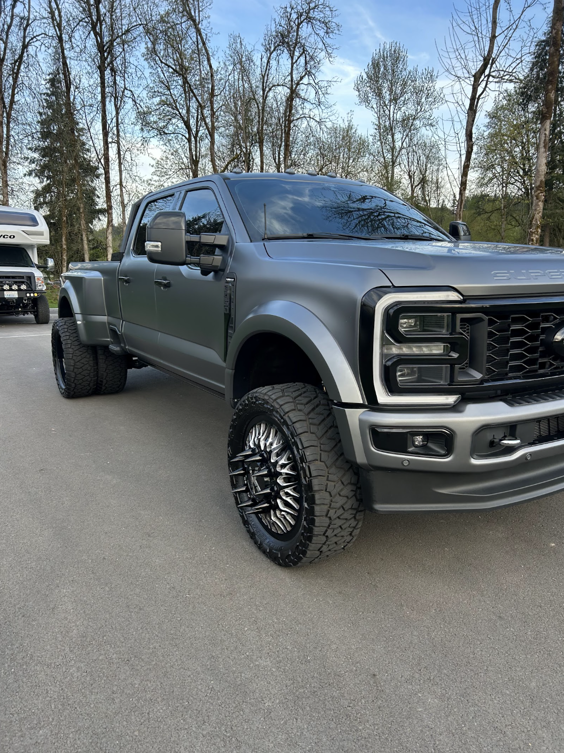 A matte gray lifted pickup truck with large off-road tires parked on a paved lot, with trees in the background and a partially visible white vehicle in the distance.