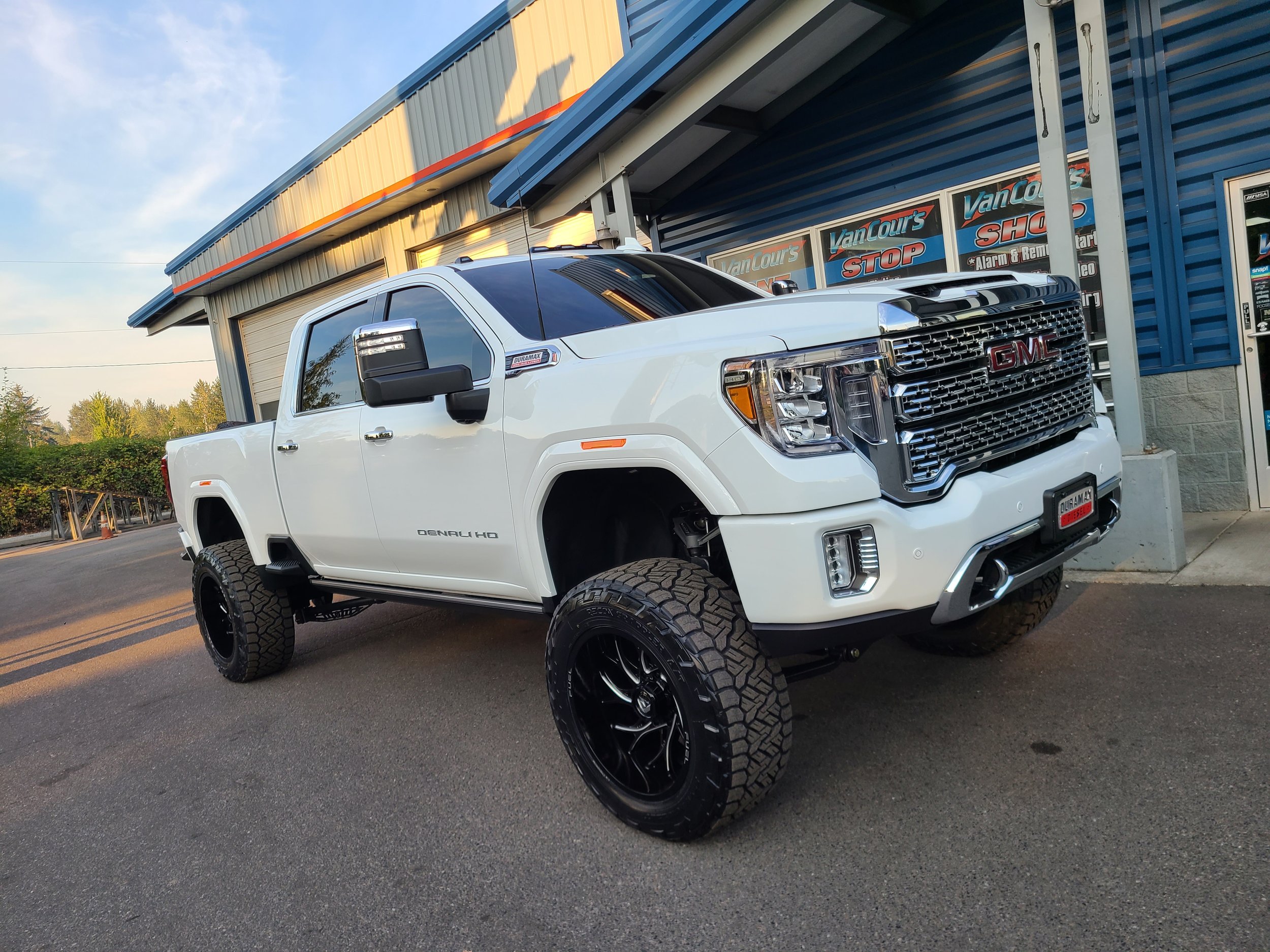A white GMC pickup truck with large black tires parked in front of a building with signs reading 'Van Cou rs STOP'. The building has a blue and gray exterior with a yellow accent stripe and a partial closed garage door. Trees and a clear sky are visi