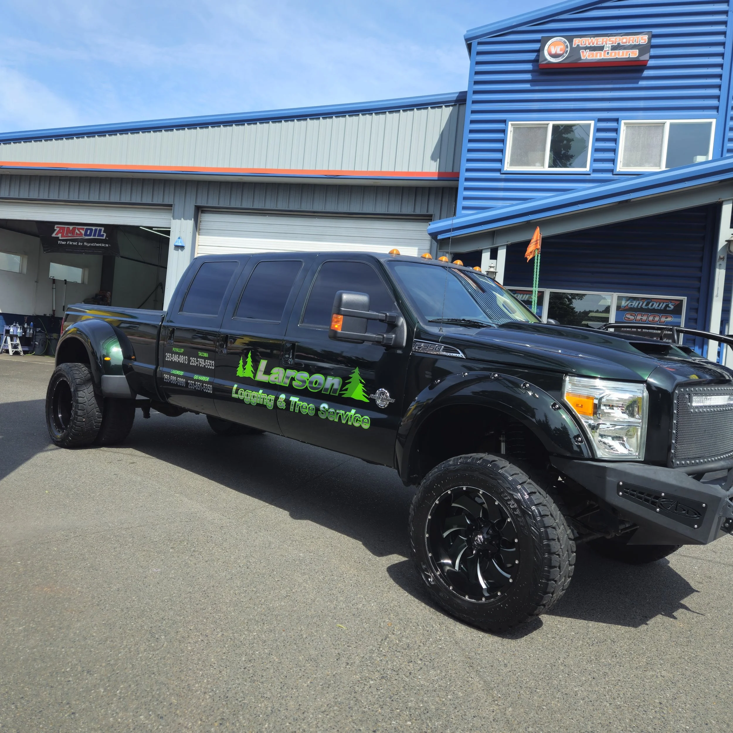 A black pickup truck with green lettering parked outside a service shop with blue and gray metal siding. The shop has signs for Powersports by VanCours and AMSOIL.