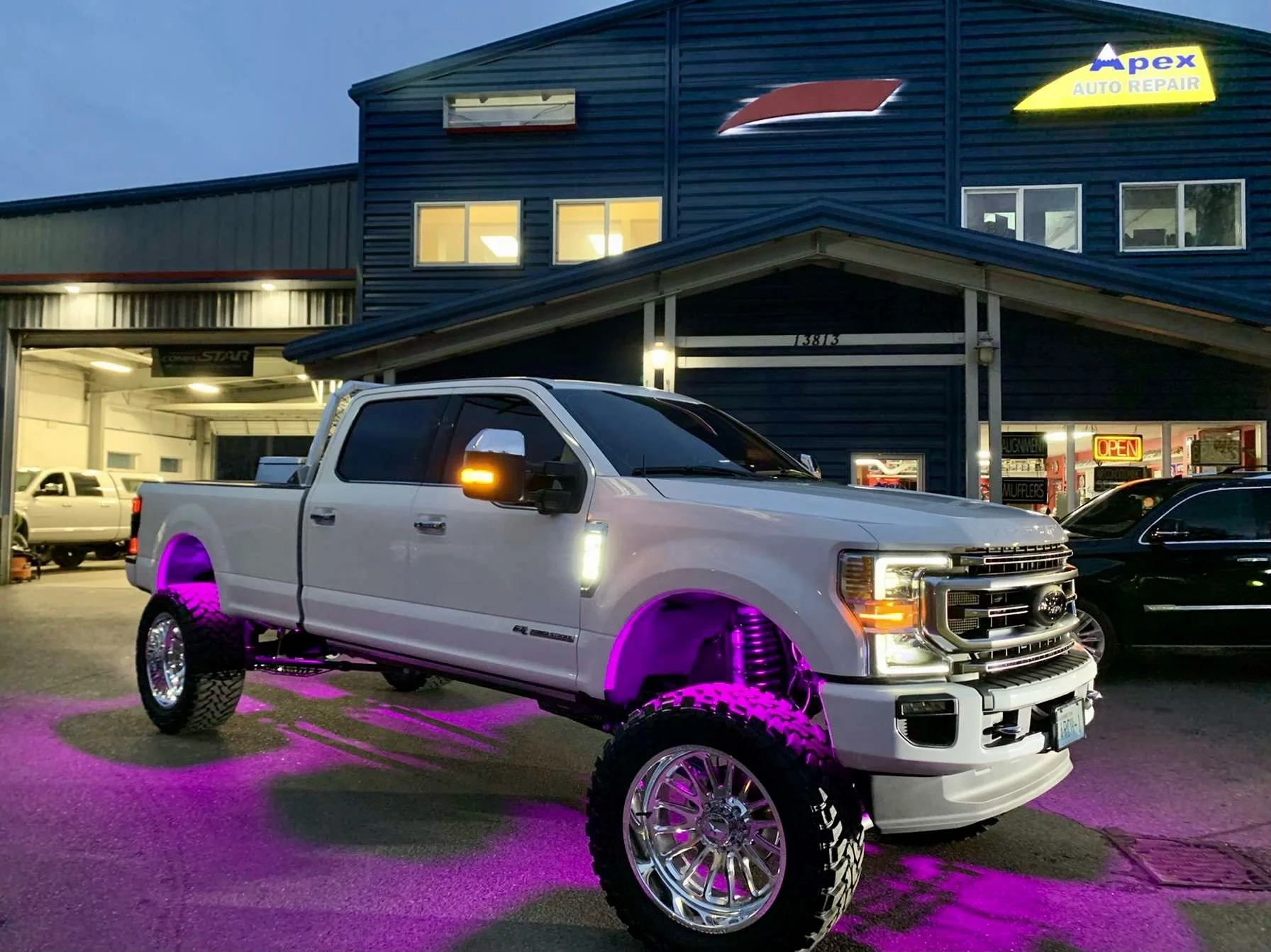 A white pickup truck with large purple underglow lights and custom oversized wheels parked outside an auto repair shop at dusk. The shop's sign reads 'Apex Auto Repair' and has a lit 'OPEN' sign. The truck is in front of a dark blue building with ill